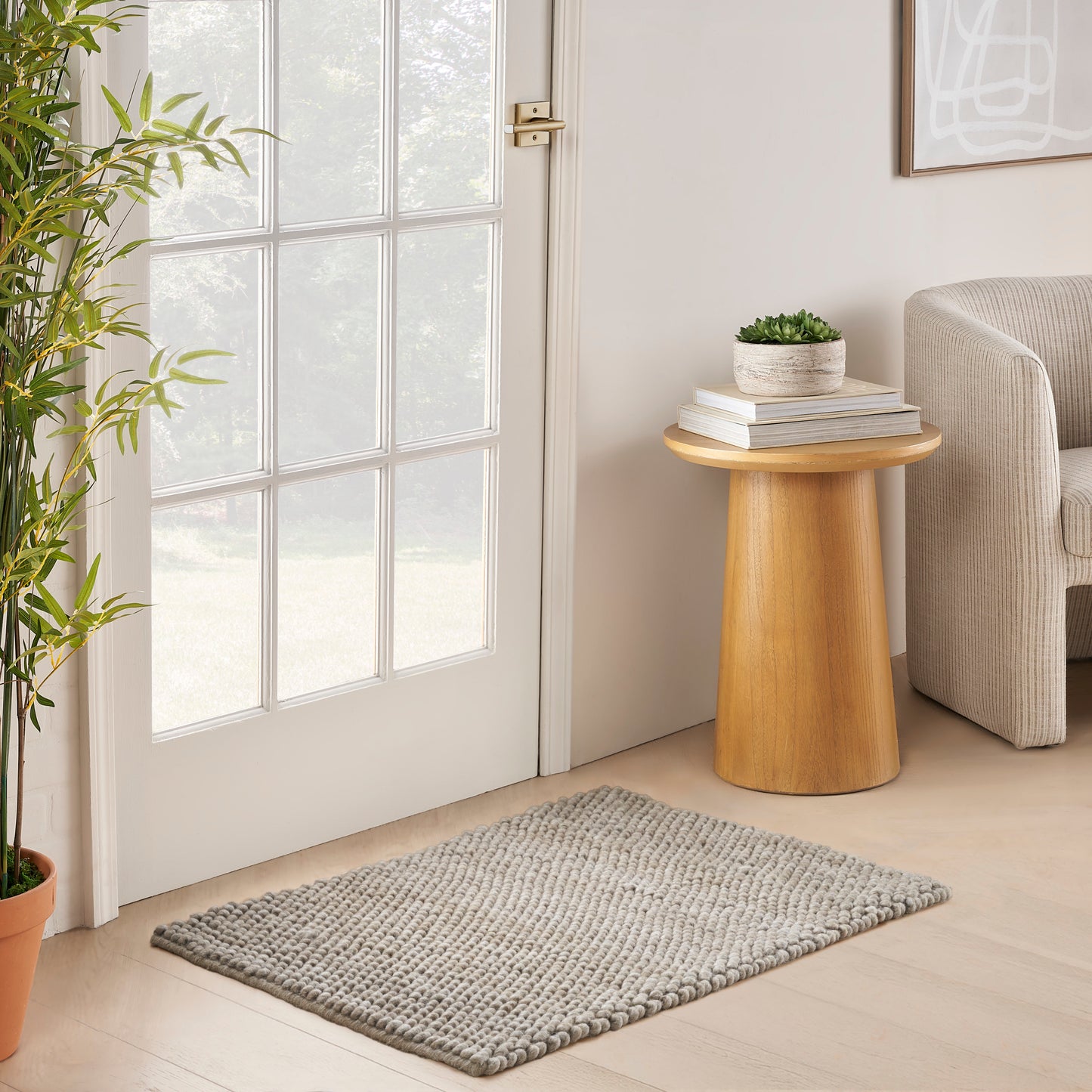 Cozy beige living nook with a glass-paned door, round wooden side table, stacked books, plant, gray rug, and a beige sofa.