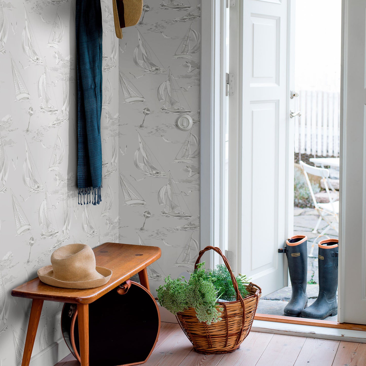 Mudroom entryway with sailboat wallpaper, wooden bench, straw hat, wicker basket with greenery, rain boots by door.