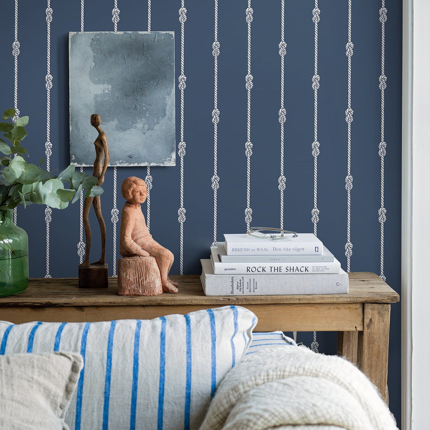 Rustic wooden console atop blue rope-pattern wall, with green vase, two wooden figures, stacked books, and blue-gray artwork.