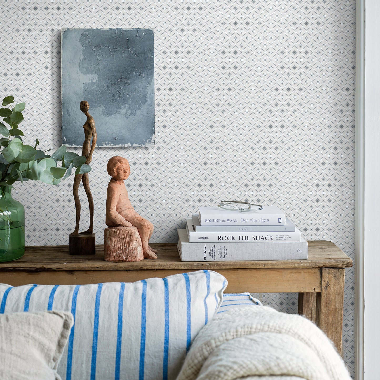 Rustic wooden console with two sculptures, stack of white books, green vase with eucalyptus, blue-gray diamond wallpaper.