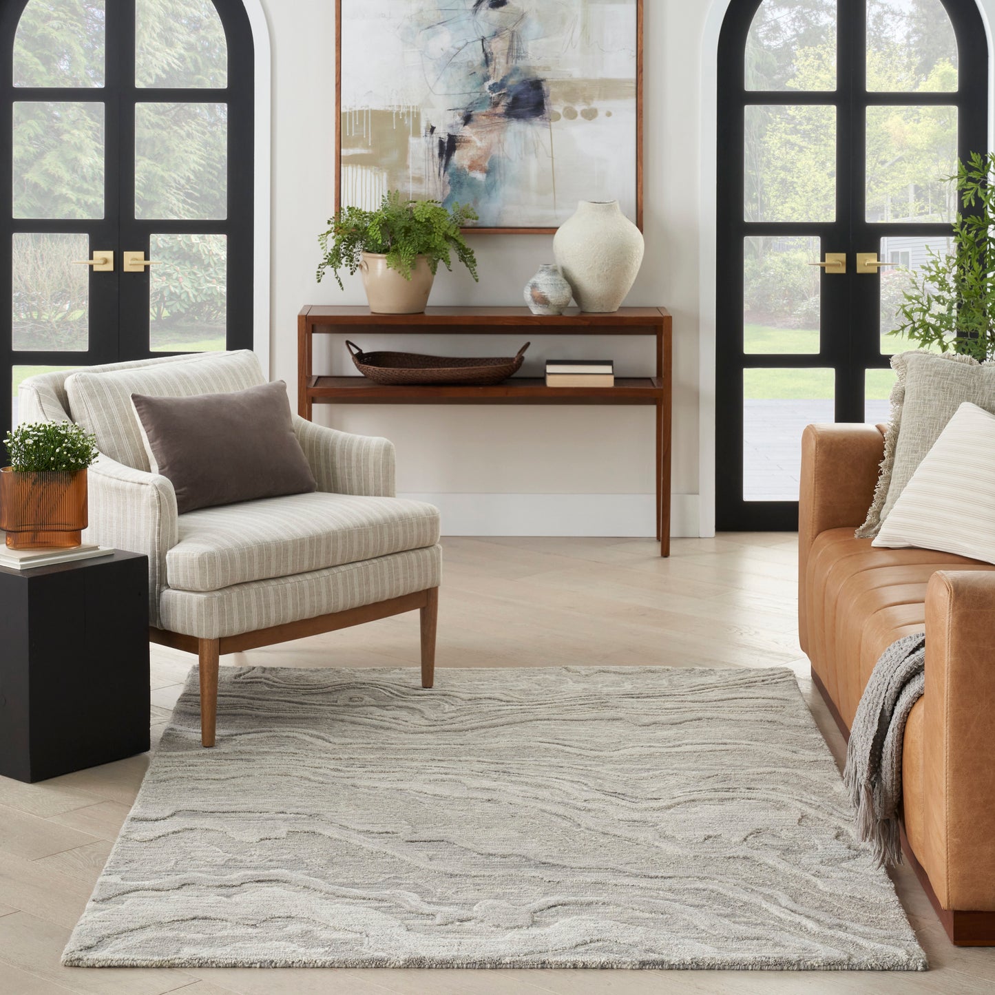 Cozy living room with beige striped armchair, tan sofa, light gray rug, console table, plants, and arched black windows.