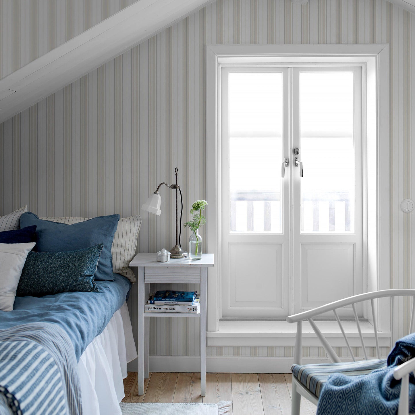 Cozy bedroom with light blue bedding, striped grey wallpaper, white bedside table with lamp, plant, and white French doors.