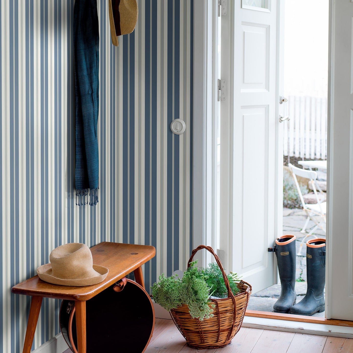 Wooden bench with straw hat, blue striped wallpaper, scarf; wicker basket with greenery and boots by the open doorway.