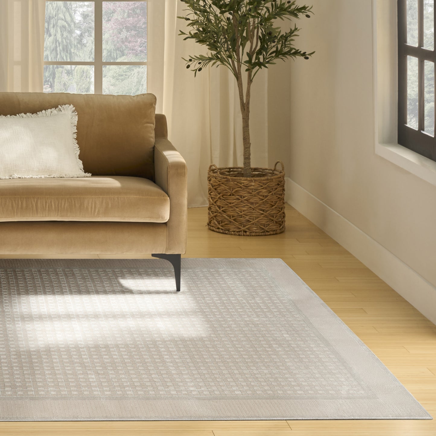 Beige sofa with white fringed pillow, sits on light geometric rug over wood floor; potted tree in woven basket nearby.