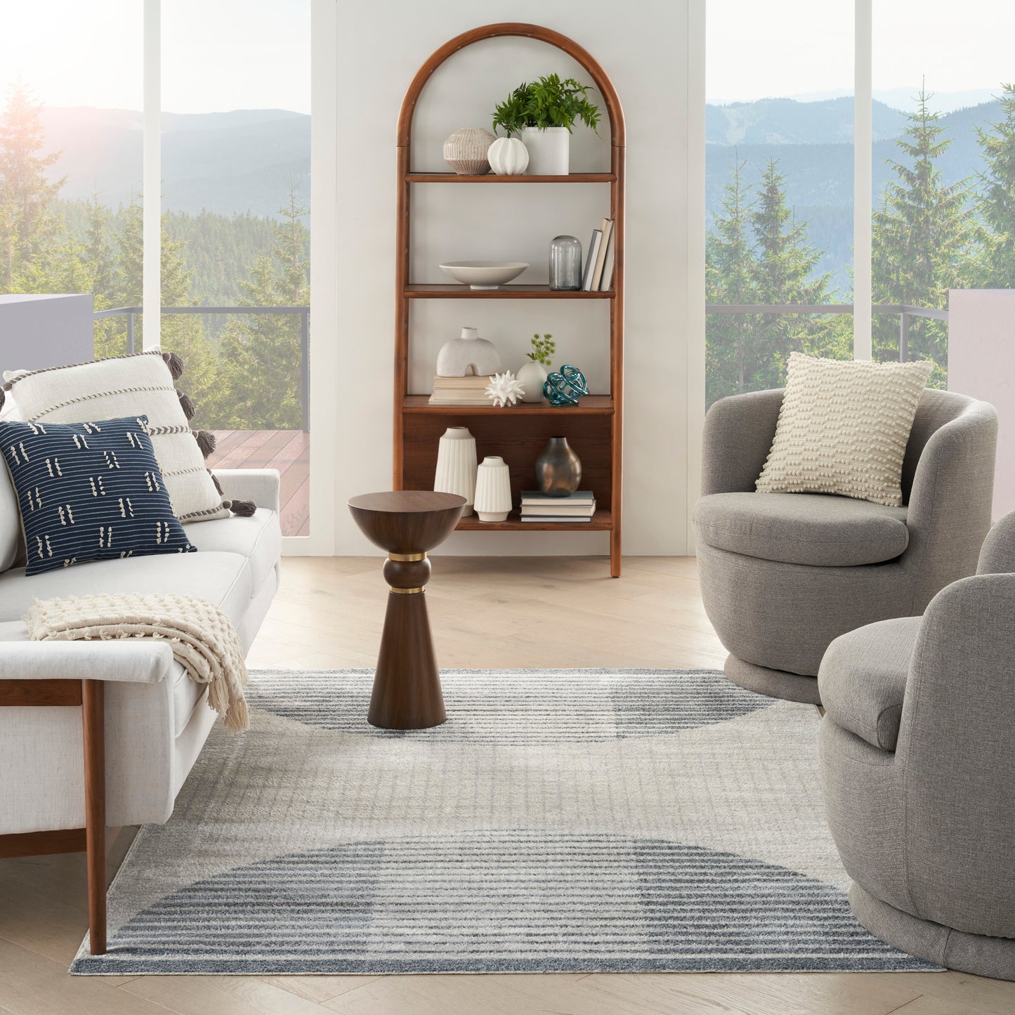Living room with an arched wooden bookshelf, white sofa, gray lounge chairs, striped blue-gray rug, and mountain view