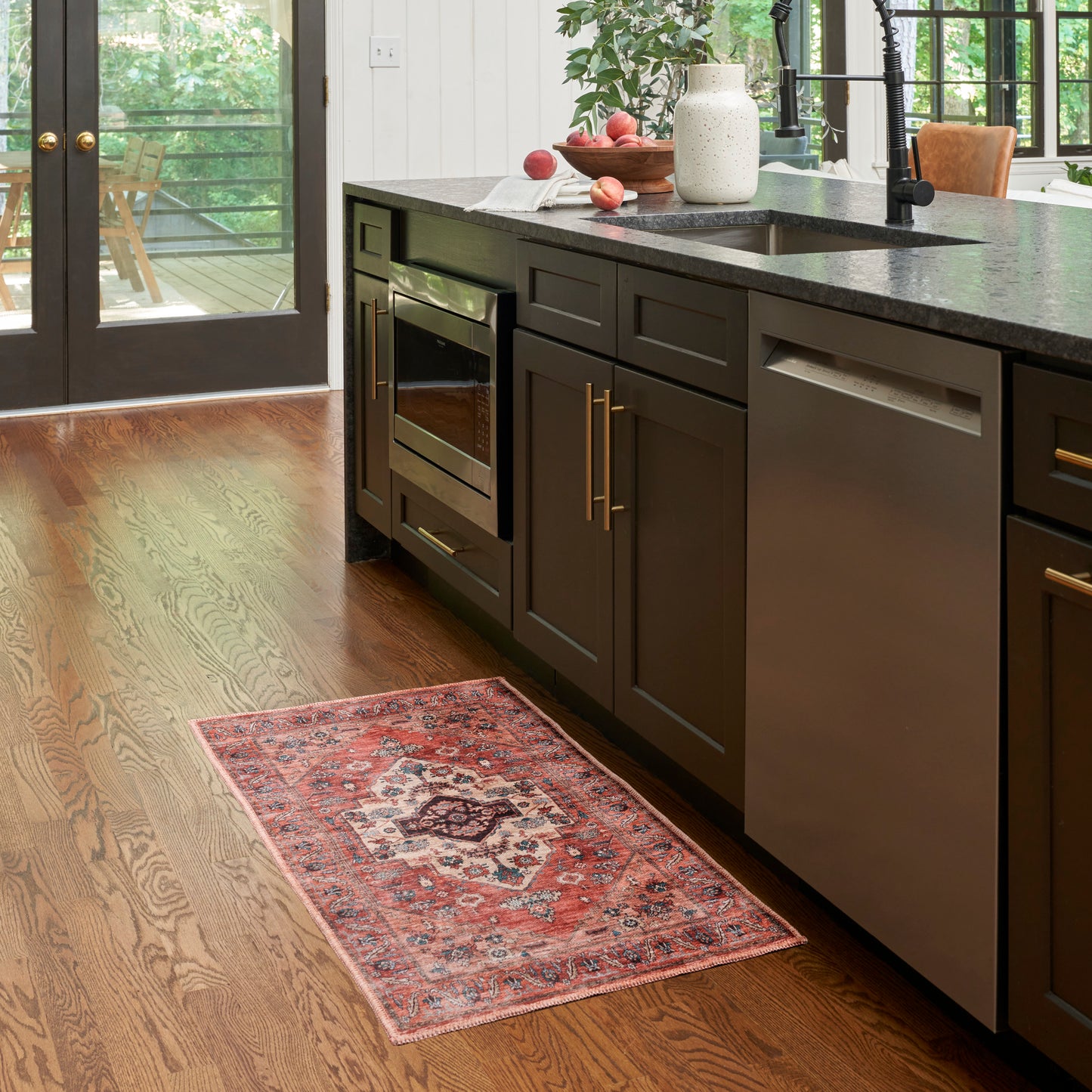 Dark wood kitchen cabinets with stainless steel oven and dishwasher, granite countertop, red patterned rug on hardwood floor.