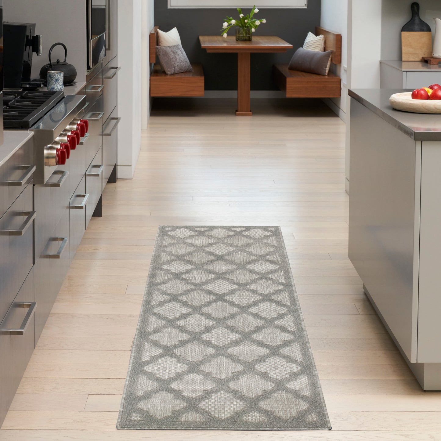 Gray lattice rug on light wood kitchen floor with stainless appliances and a cozy dining nook in back.