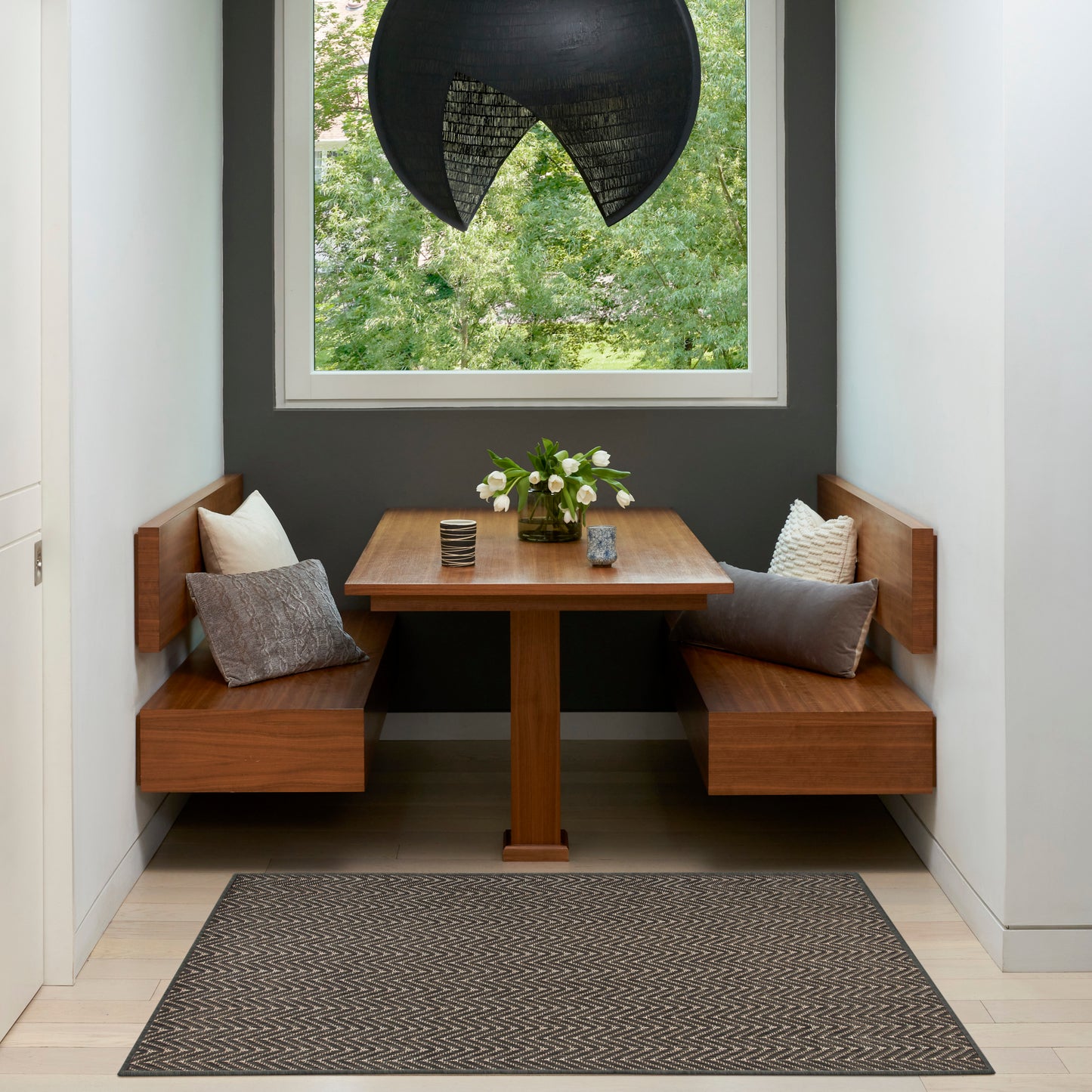 Warm wooden built-in dining nook with square table, grey cushions, and a chevron rug under a vase of white flowers.