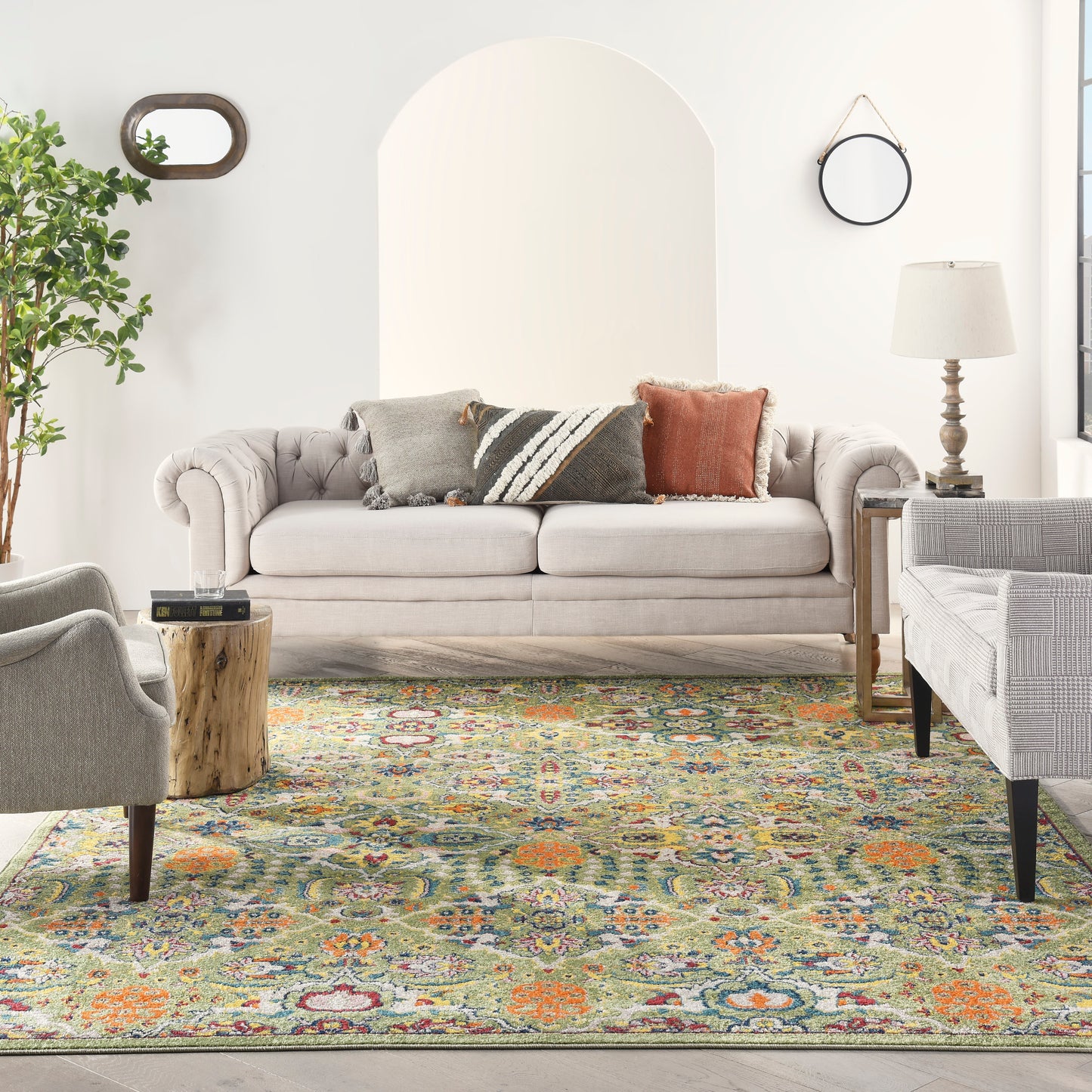 Beige tufted sofa with gray and rust cushions, multicolor oriental rug, arched wall niche, plant, round mirror, and lamp.