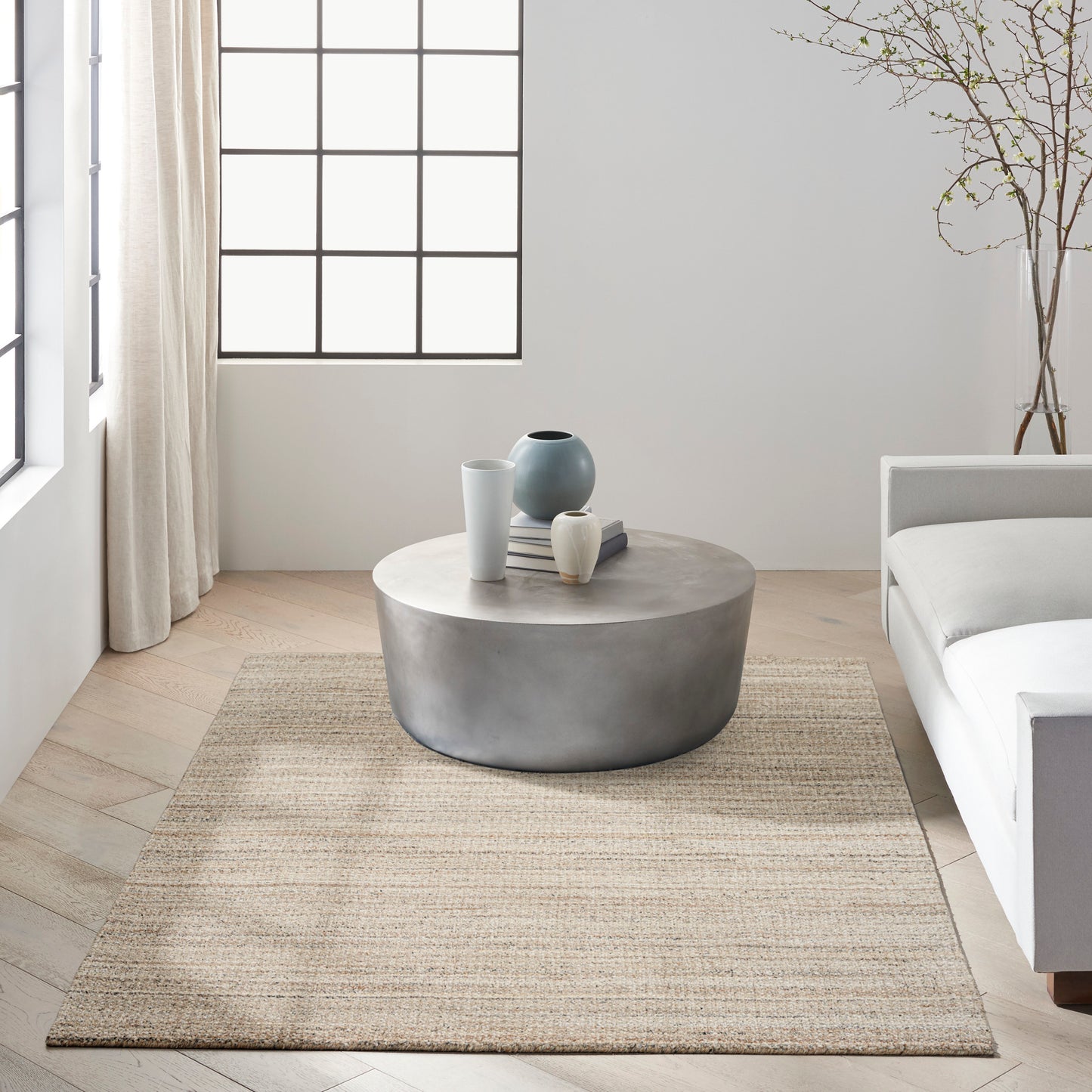 Beige woven rug in a bright minimalist living room, with a round coffee table, ceramic vases and books visible.