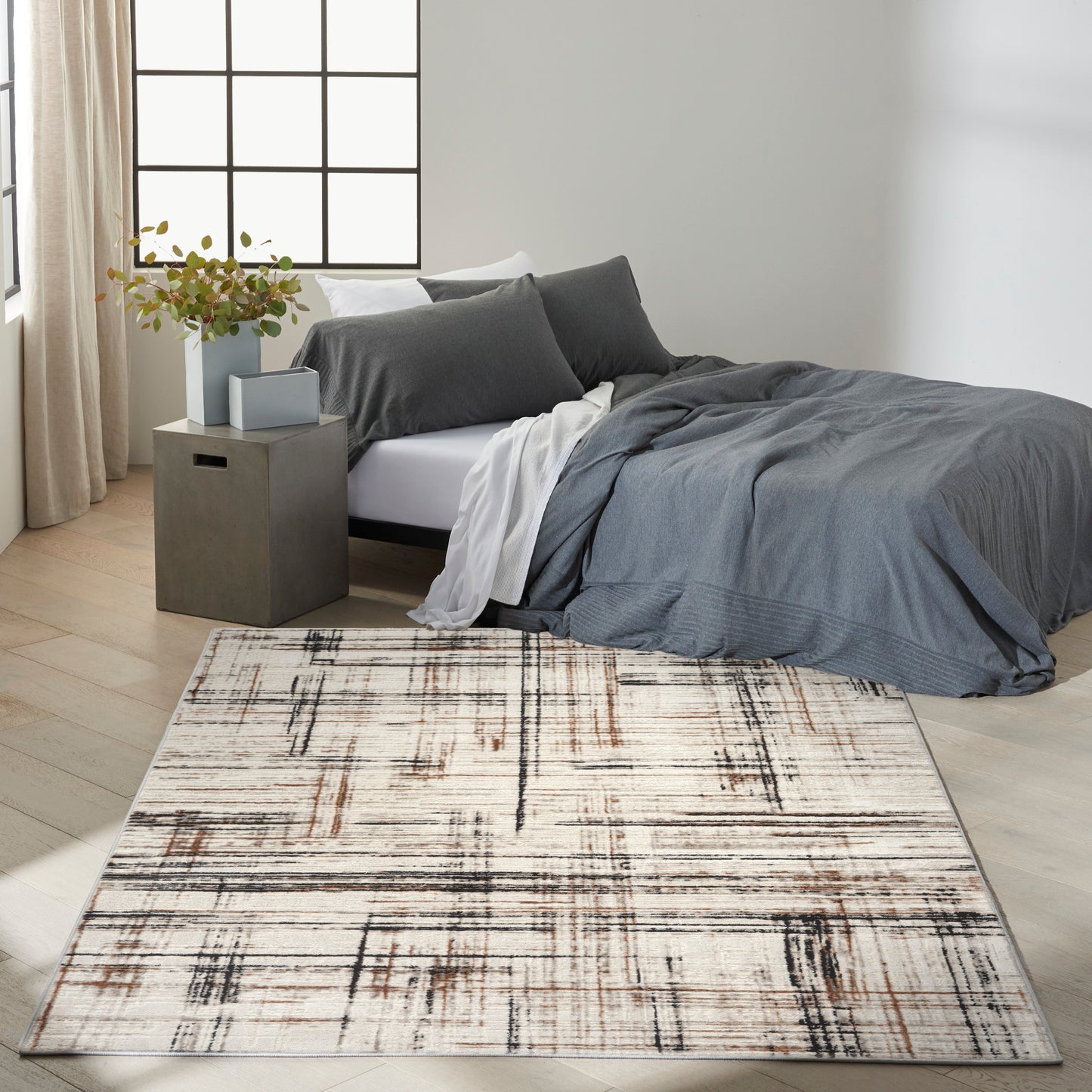 Modern bedroom with charcoal gray duvet, matching pillows, beige-and-brown abstract rug, concrete side table, potted plant.