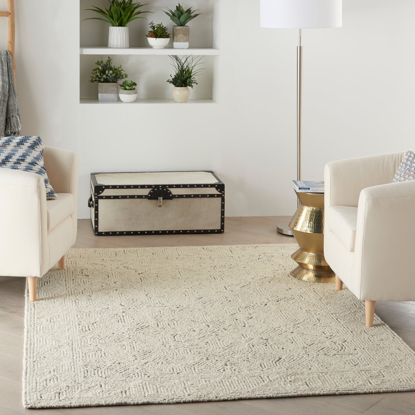 Cream upholstered chairs, beige woven rug, vintage studded trunk, brass side table, and potted plants in a living room.