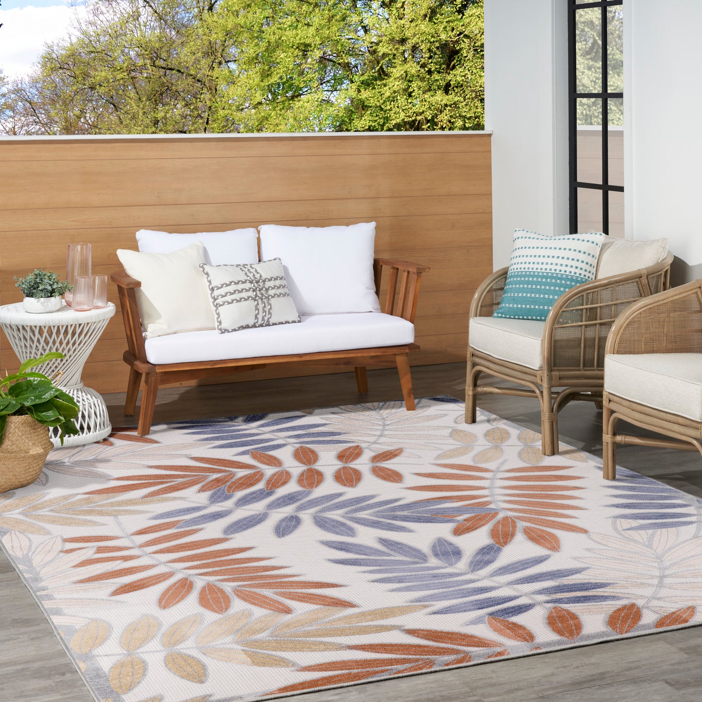 Outdoor seating with white cushioned wooden sofa, wicker chairs, potted plants, glass side table, and orange-blue leaf rug.