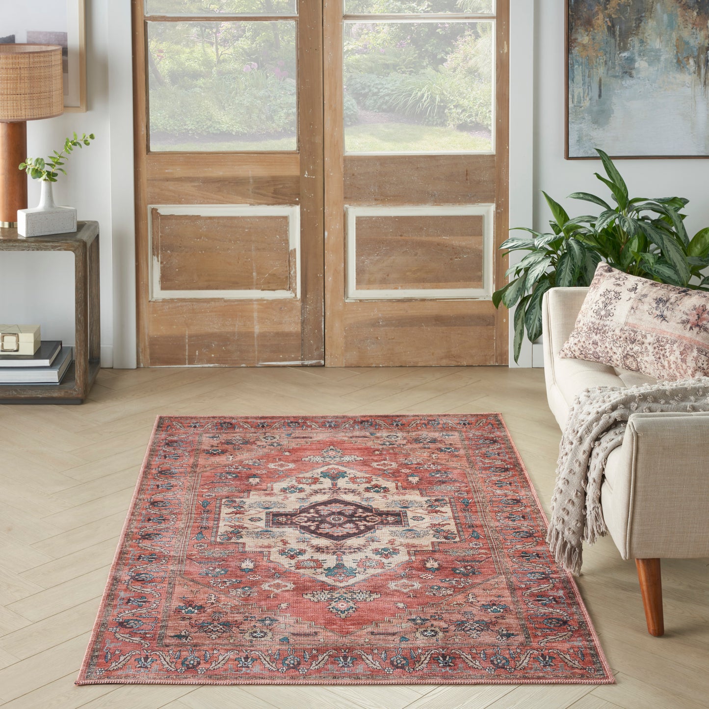 Persian-style red area rug with blue and ivory borders on light wood floor, beside beige sofa and houseplant.