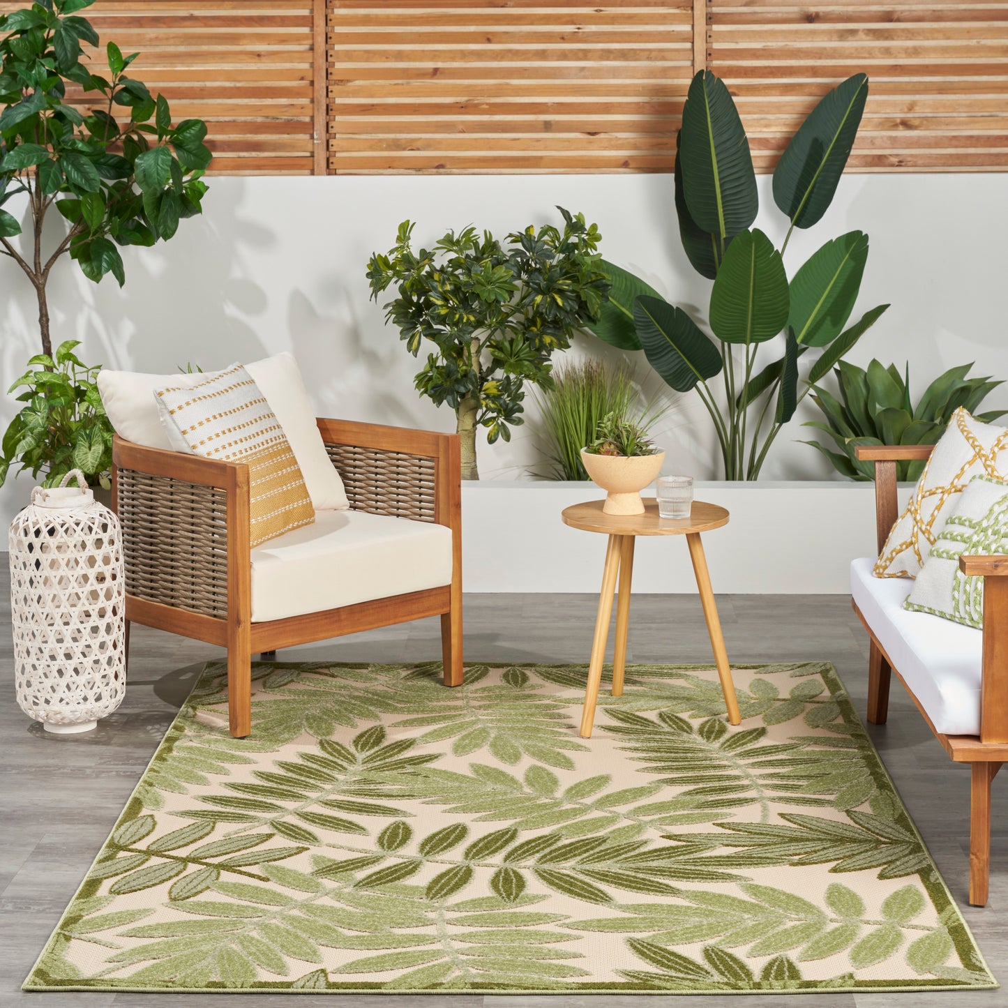Wooden-frame seating with beige cushions, round side table, & a green leaf-pattern rug amid potted tropical plants.