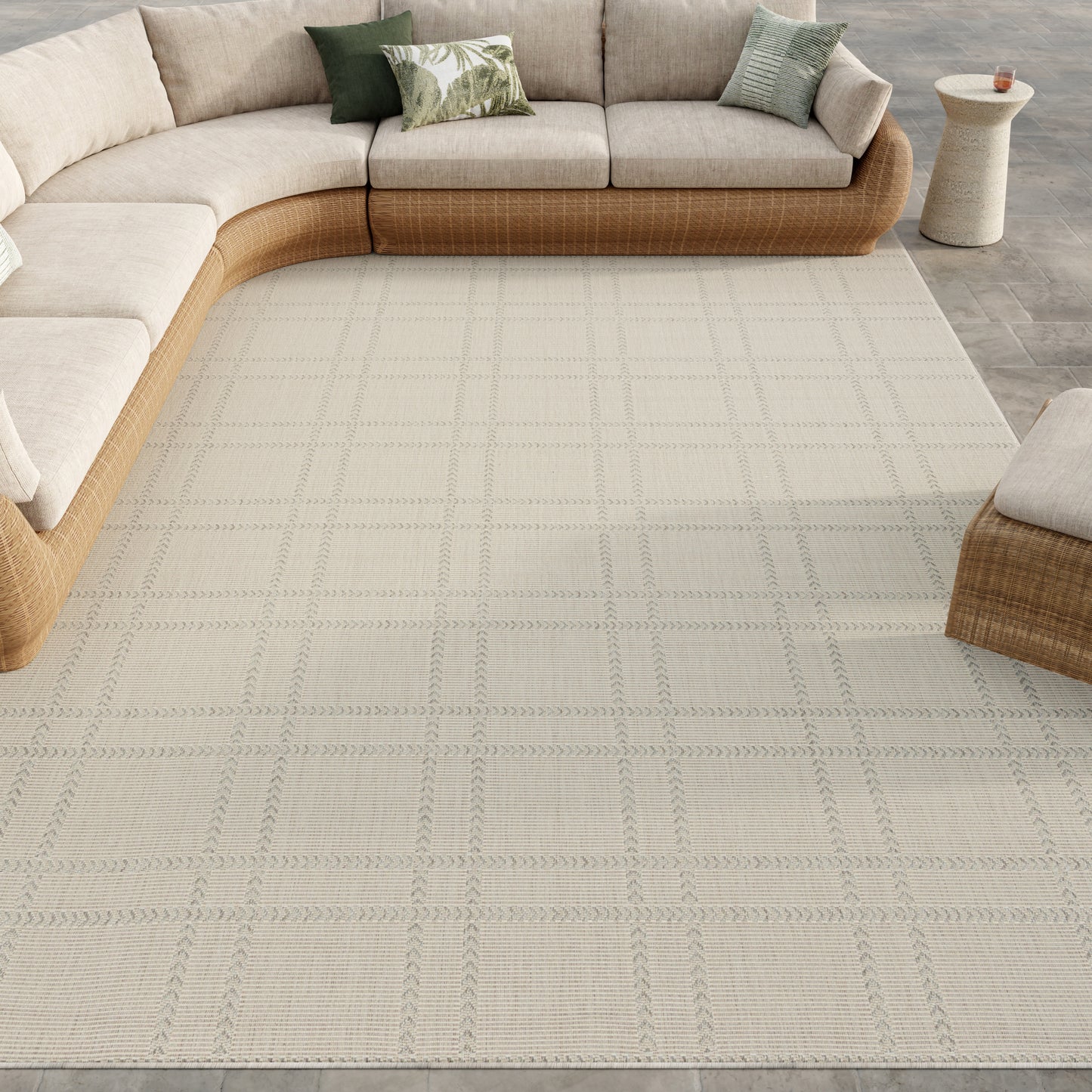 Beige sectional with woven wicker base, light grid-pattern rug, tropical green pillows, and a small beige patio side table.