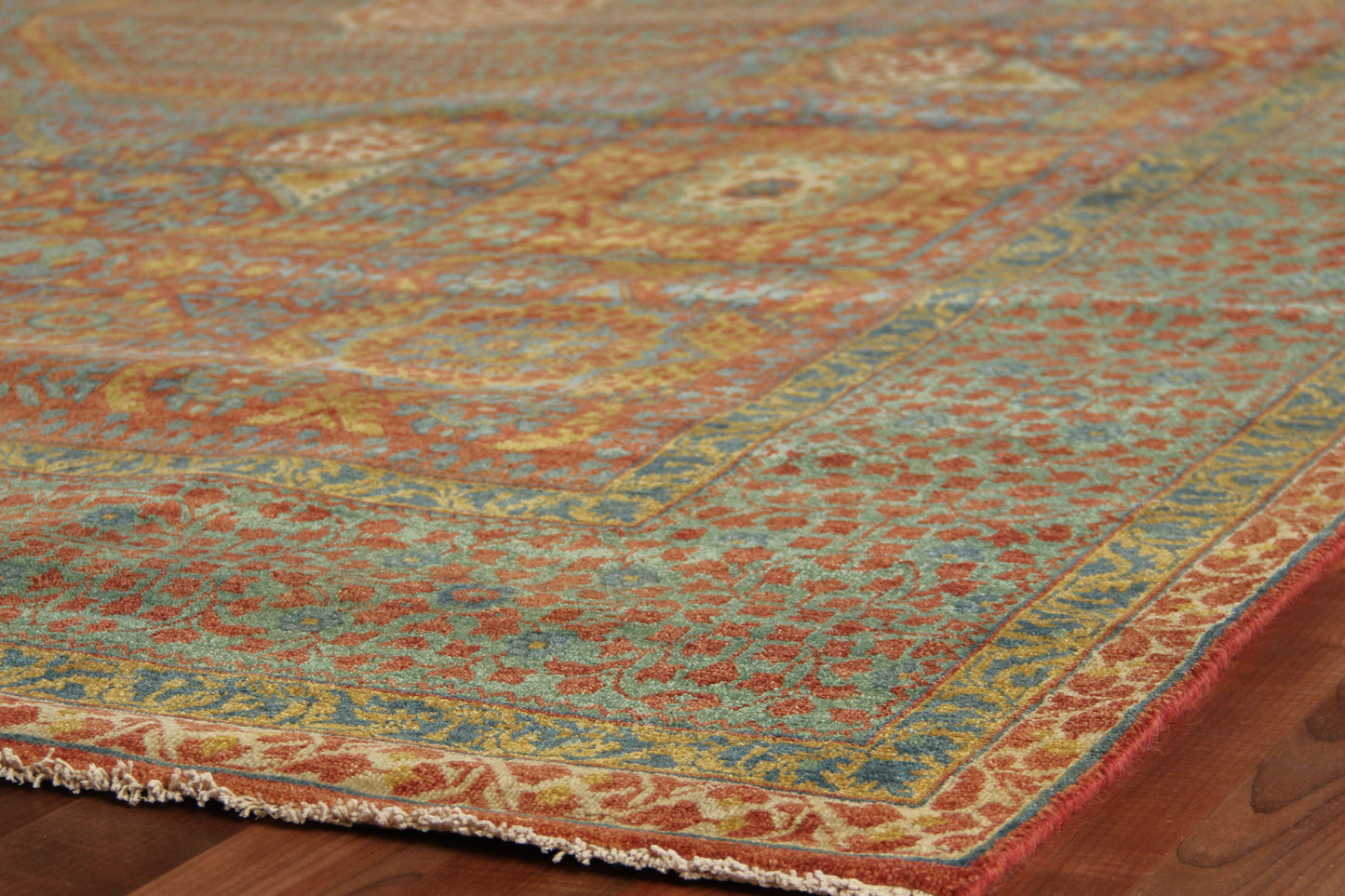 Traditional oriental rug on a wood floor with teal, orange & gold tones, an intricate floral border and fringed edges.