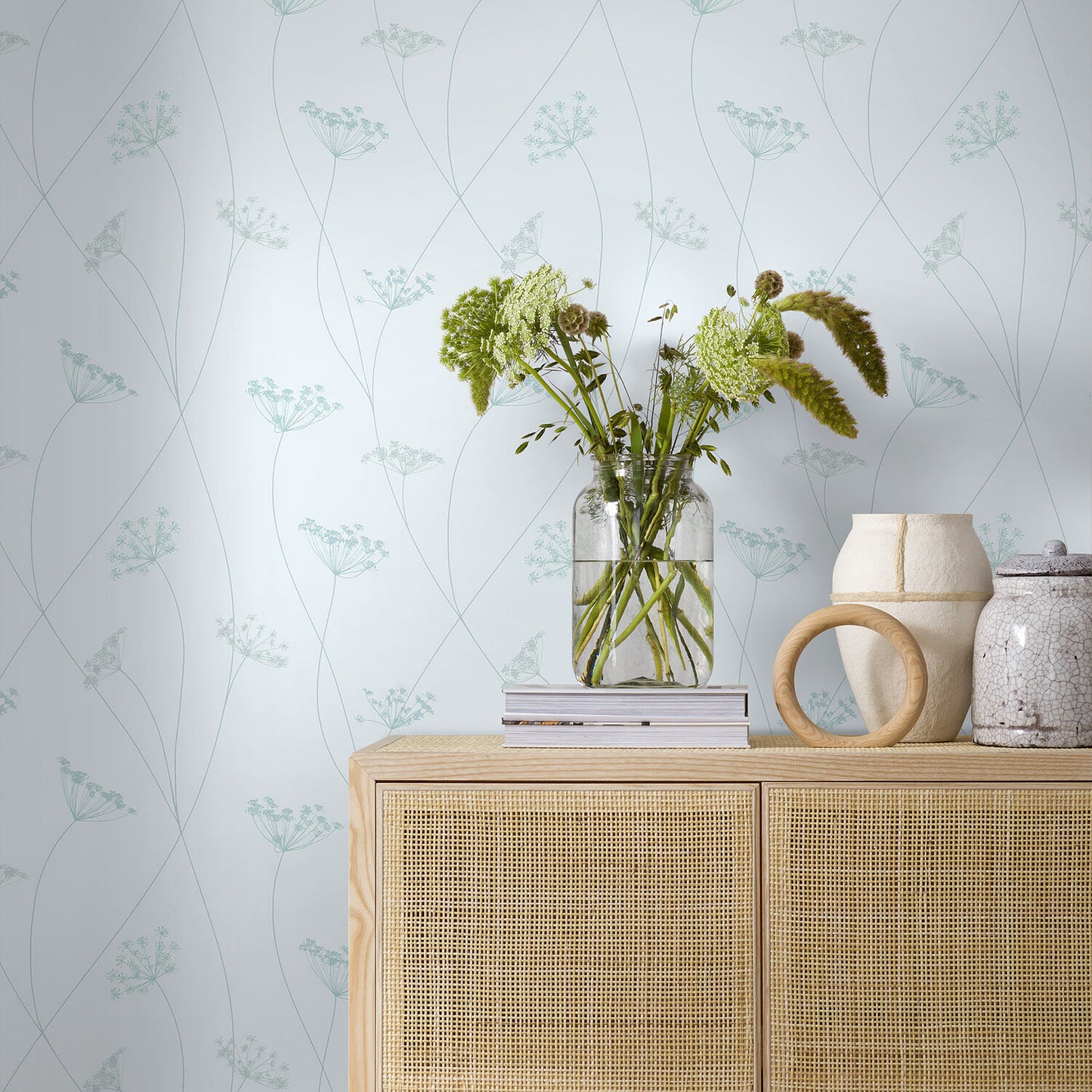 Light wood sideboard with cane doors, glass vase of flowers, books, and ceramic vases on pale botanical wallpaper.