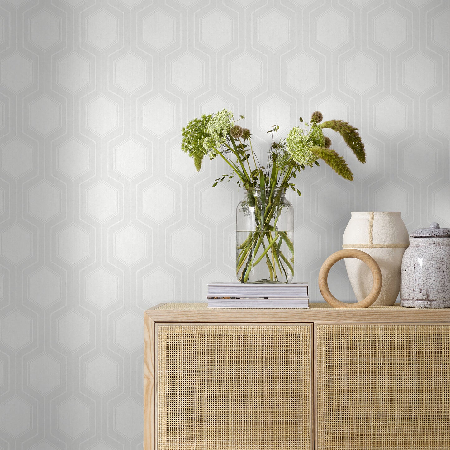 Light wood sideboard with cane panels, glass vase of greenery, stacked books, and ceramic jugs against pale hex wallpaper.