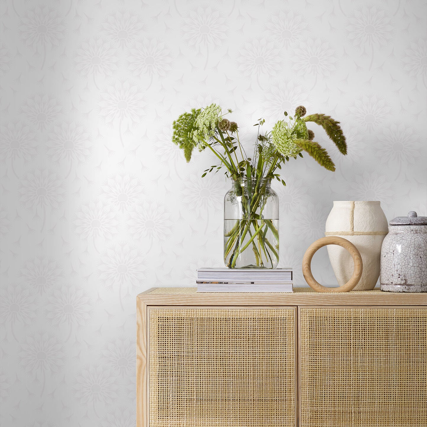 Light wood sideboard with rattan-front panels, glass vase of green stems, stack of books, and ceramic vessels.