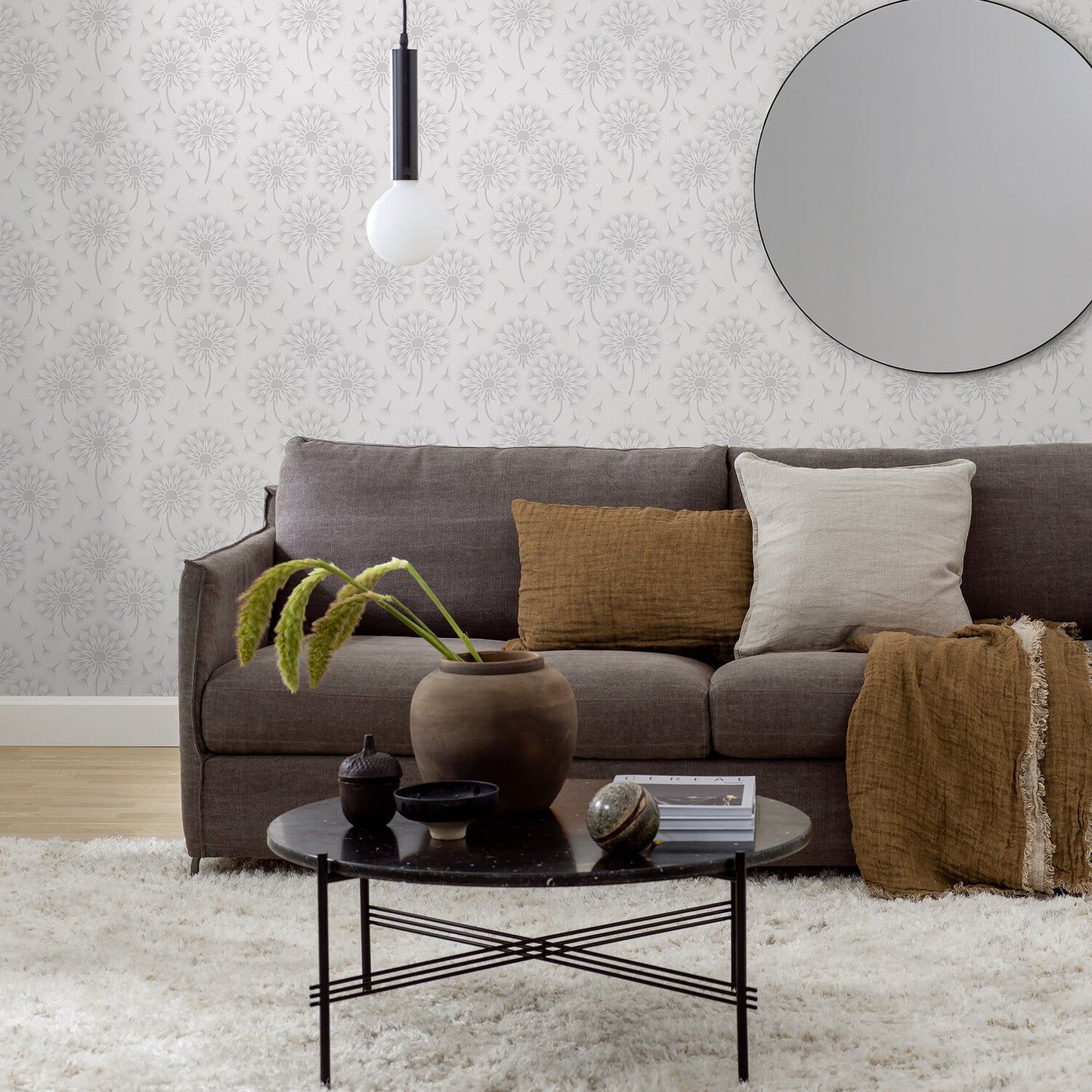 Grey sofa with brown and beige cushions and a rust throw; round black coffee table with a plant, bowls, and a book; patterned wallpaper, round mirror, and a hanging globe light.