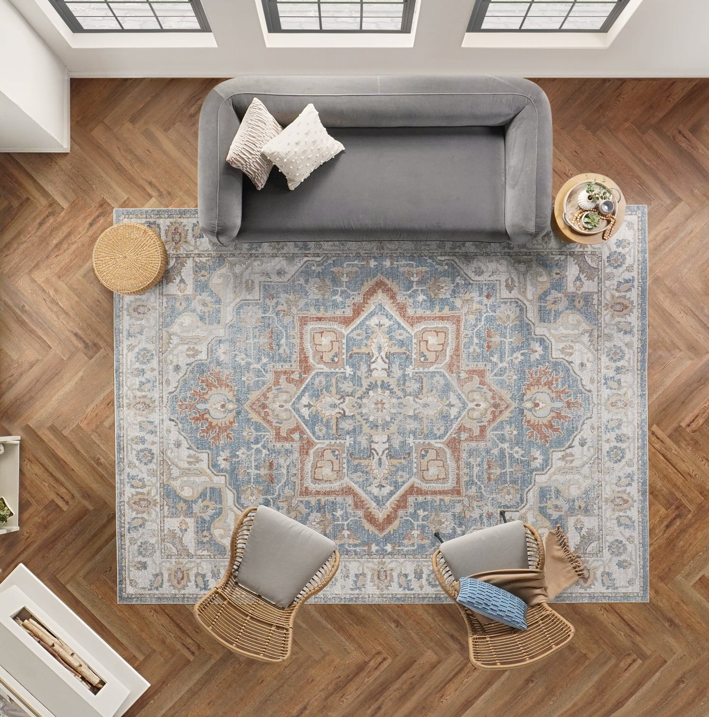 Overhead view: blue/peach patterned rug on herringbone floor, gray sofa, two wicker chairs, round ottoman, plant table.