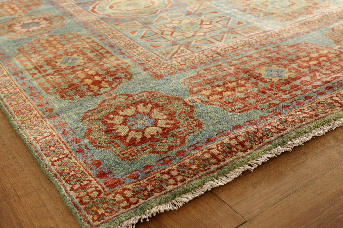 Ornate oriental rug with red, blue & teal tones, geometric medallions, floral border & fringe on a wooden floor.