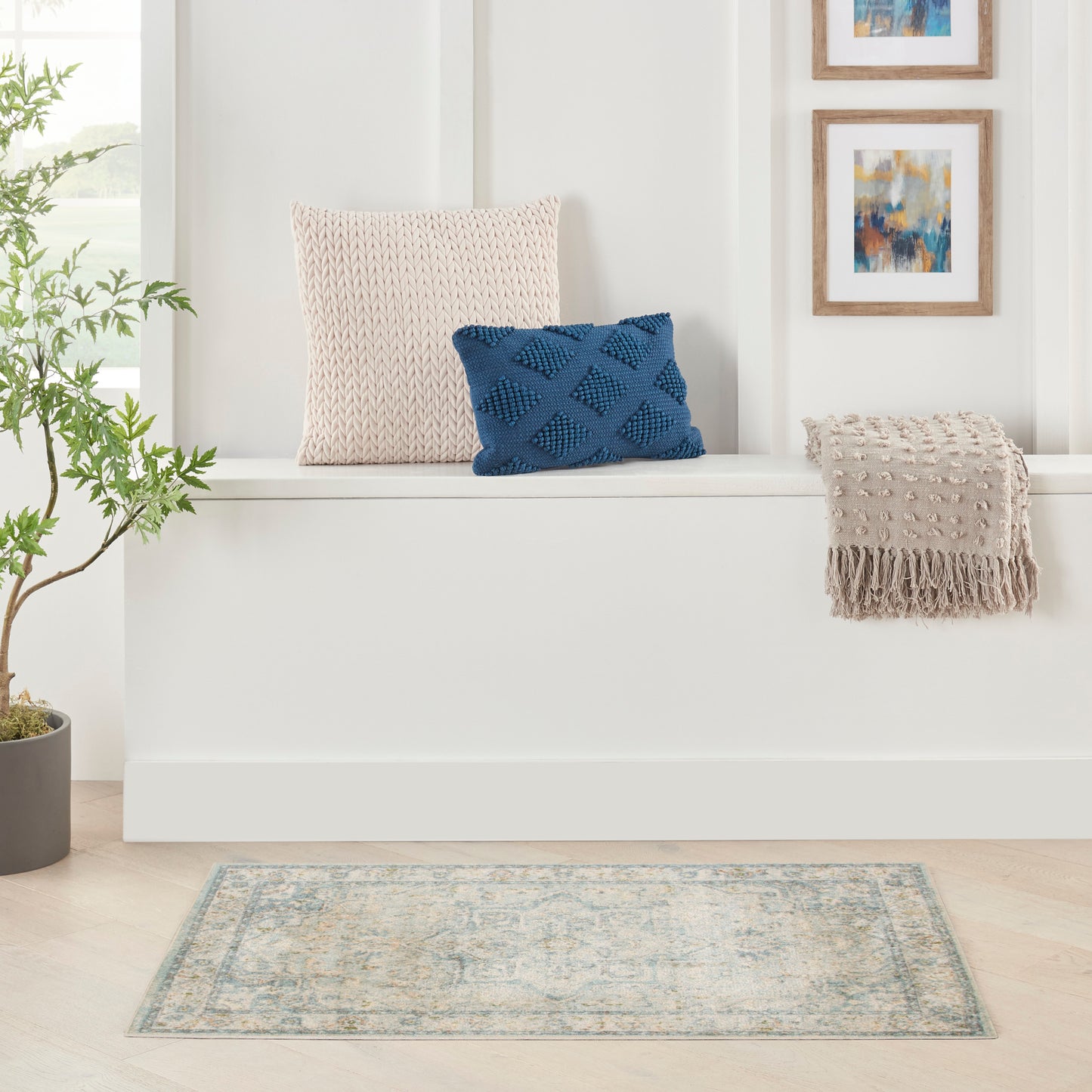 Beige knitted square cushion, navy textured diamond pillow, beige fringe-throw on white bench; blue rug, greenery, art.