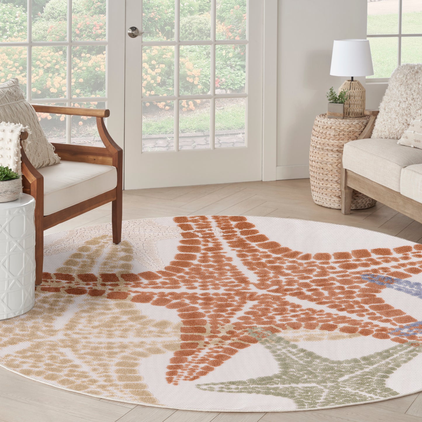 Round coastal rug with orange starfish motif and tan accents in a light living room with wicker table and cream seating.