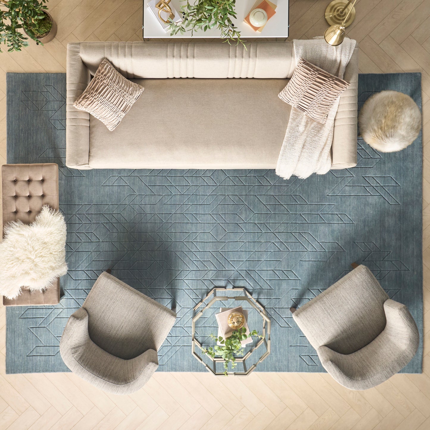 Top-down living room with beige sofa and chairs around a hexagonal glass coffee table on a blue geometric rug, with plants and throws.