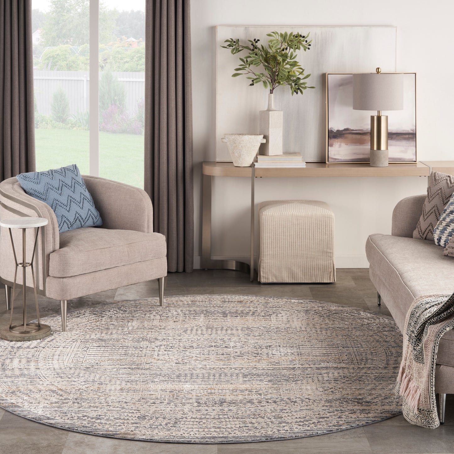 Neutral living room with round patterned rug, beige chairs, blue chevron pillow, console table, plant, lamp, and ottoman.