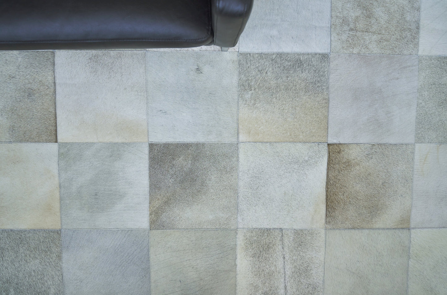 Patchwork square cowhide rug tiles in gray, beige and taupe tones with a black leather sofa edge visible.