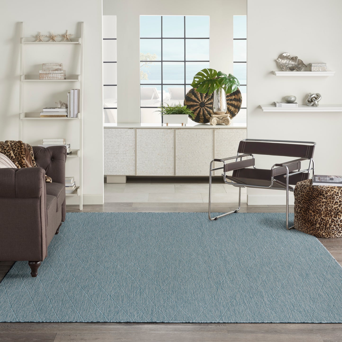 Modern living room with a light blue diamond rug, brown leather sofa, metal lounge chair, leopard ottoman, white shelves.