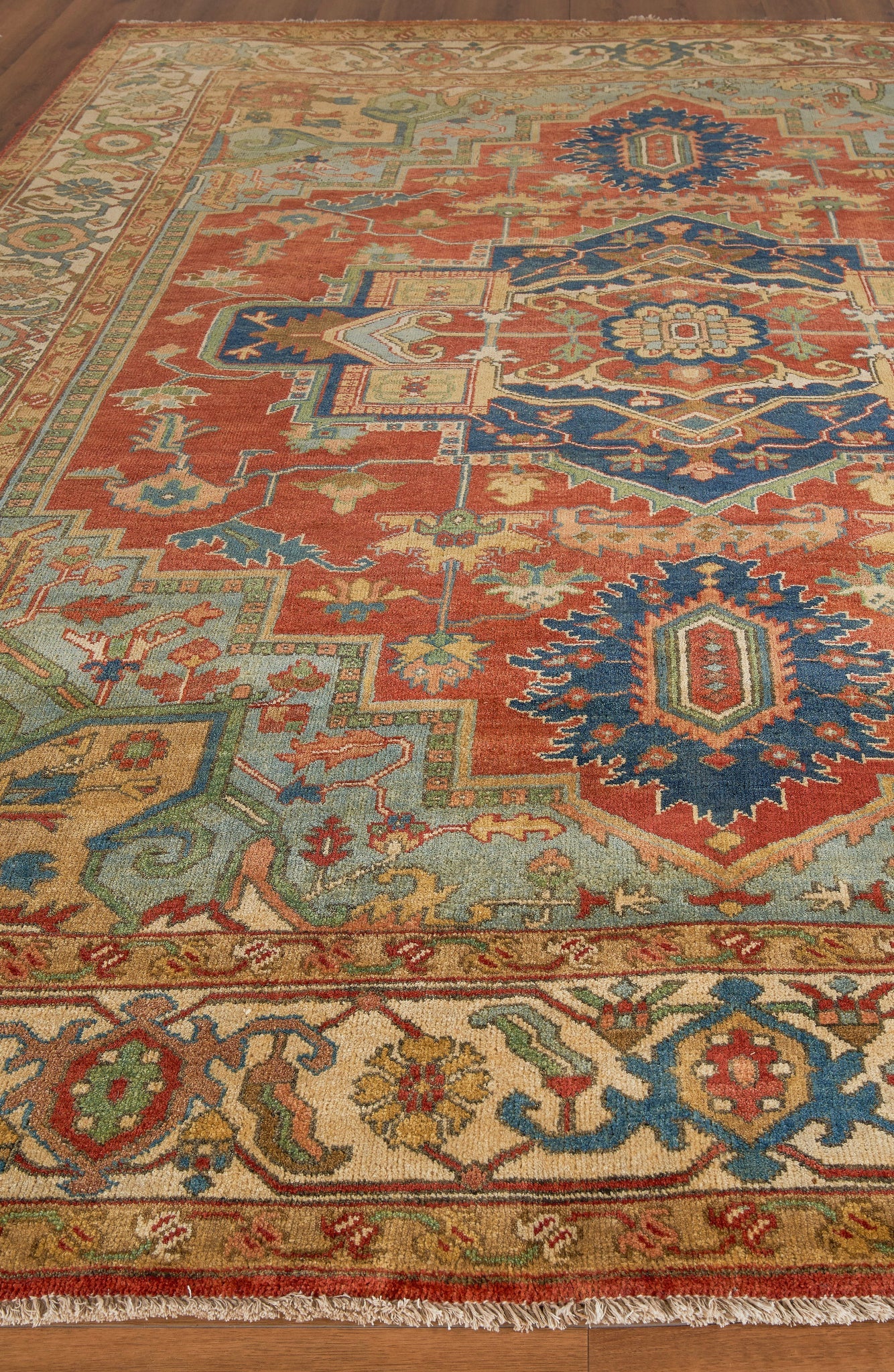 Colorful oriental-style rug with red field, blue medallions, ivory/green accents and ornate border on wood floor.