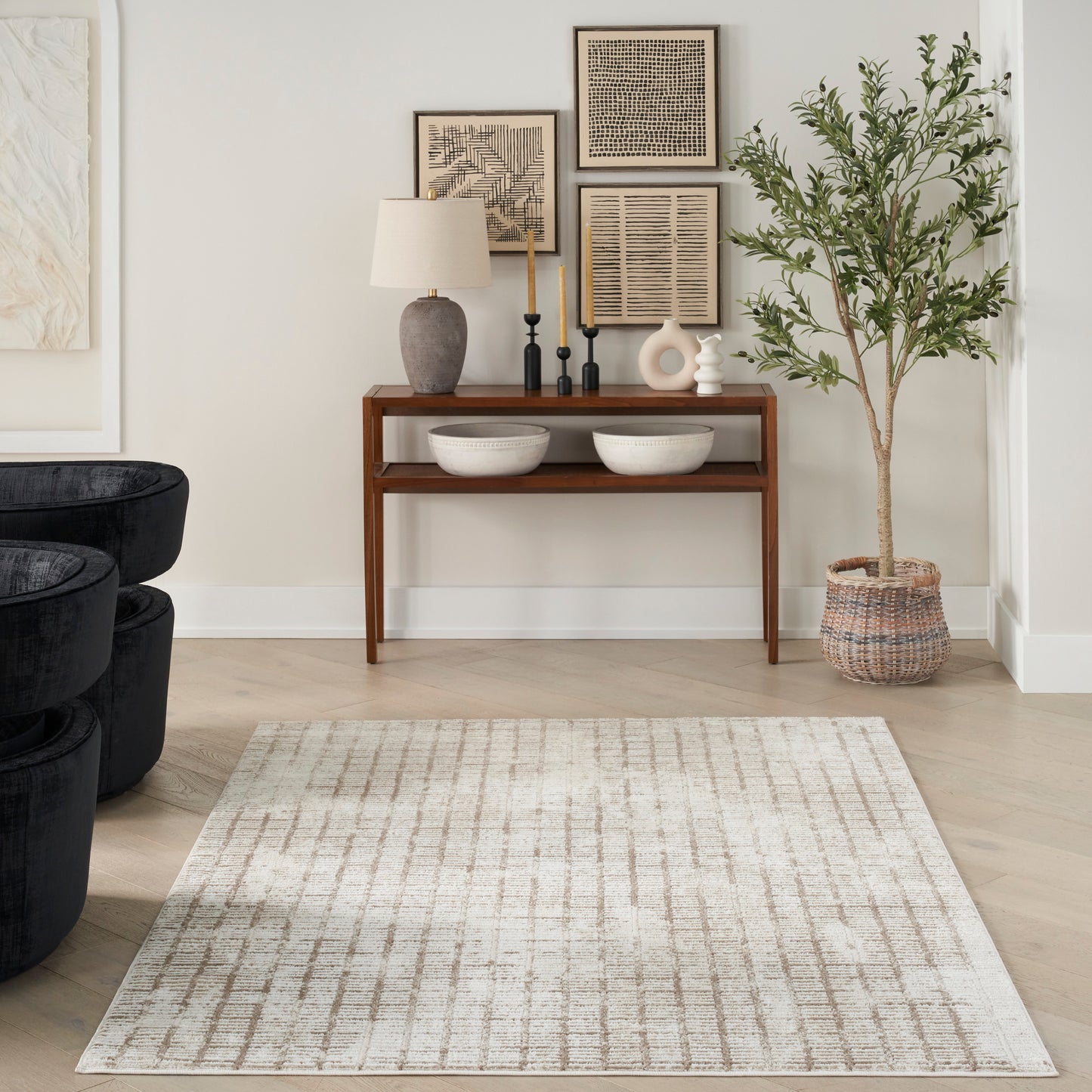 Neutral living room with an 8' x 10' rug, dark wood console, ceramic lamp, bowls, black chairs, framed art, and a tall plant in a woven basket.