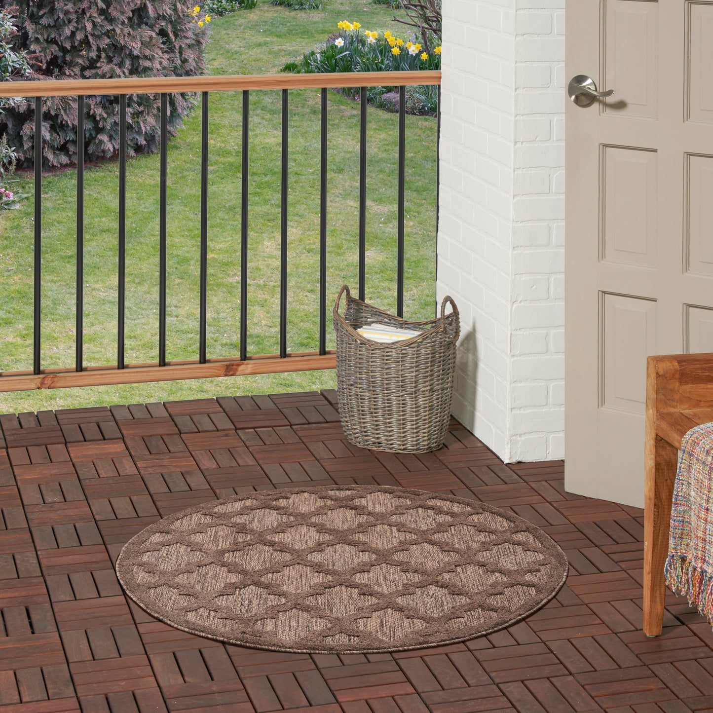 Round brown braided outdoor rug on a wooden deck beside a wicker basket and doorway.