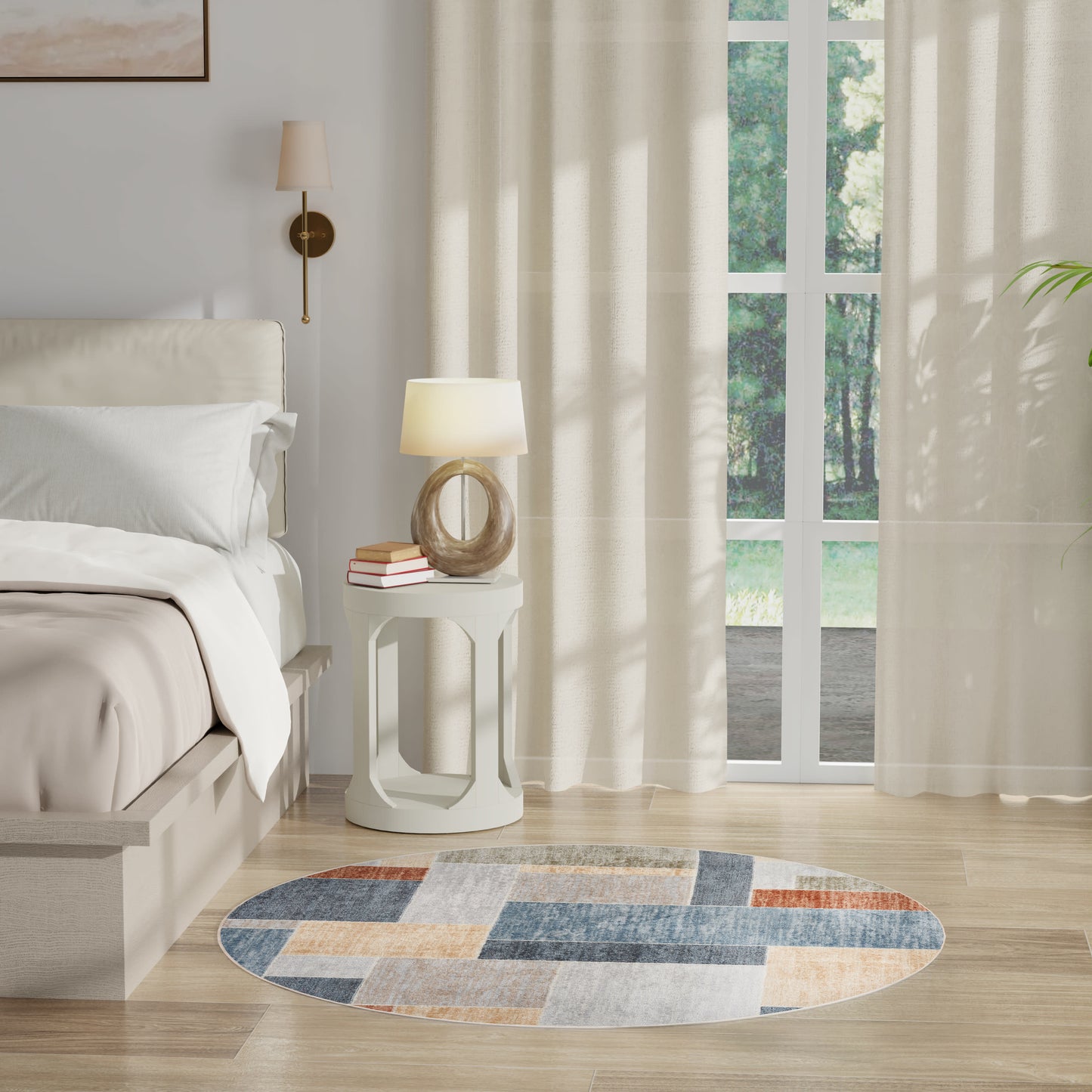 Cream bed with white linens, round white side table with lamp and books, patchwork round rug, sheer curtains, glass doors.