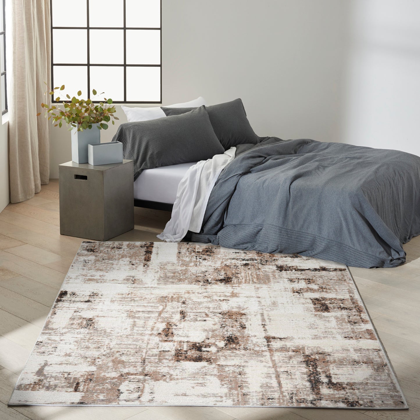 Cozy modern bedroom with charcoal gray duvet, white sheets, concrete nightstand, potted plant, and abstract cream-brown rug.