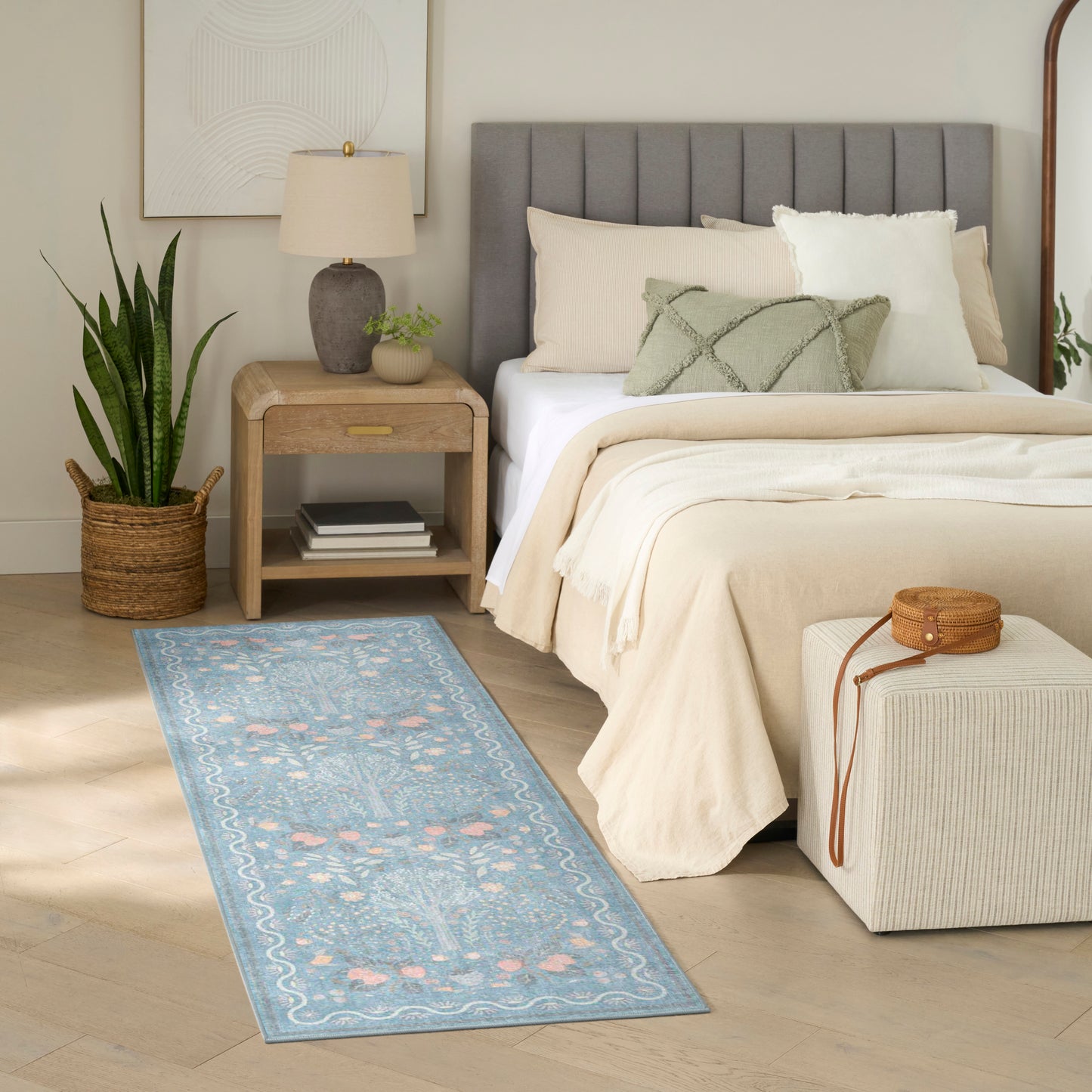Neutral bedroom featuring a gray upholstered headboard, beige bedding, blue floral runner rug, wooden nightstand, snake plant, and an ottoman.