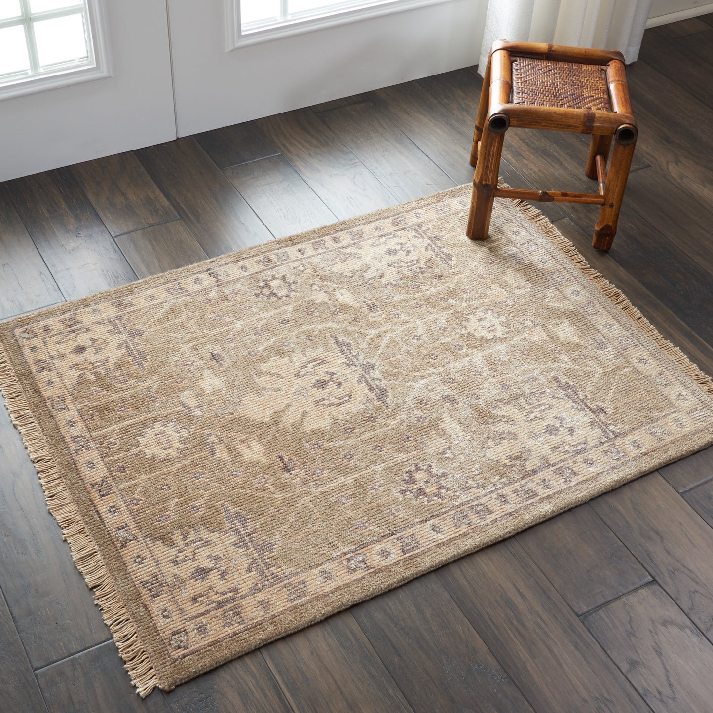 Rectangular beige woven rug with fringe and faded oriental motif on dark wood floor; small rattan stool nearby.