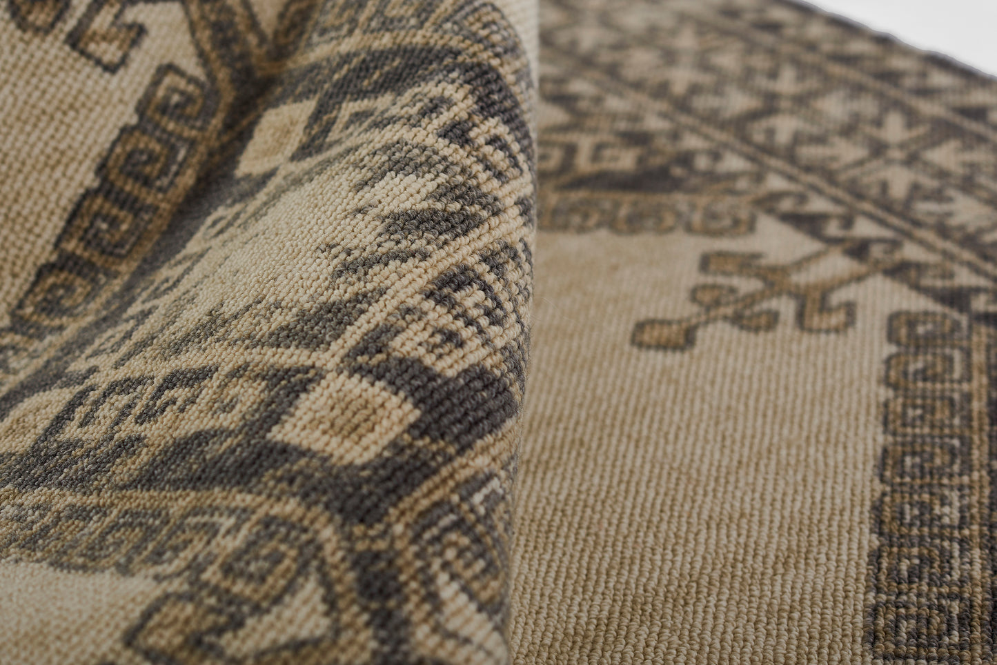 Beige and brown woven oriental rug with geometric motifs and decorative border; rolled corner visible.