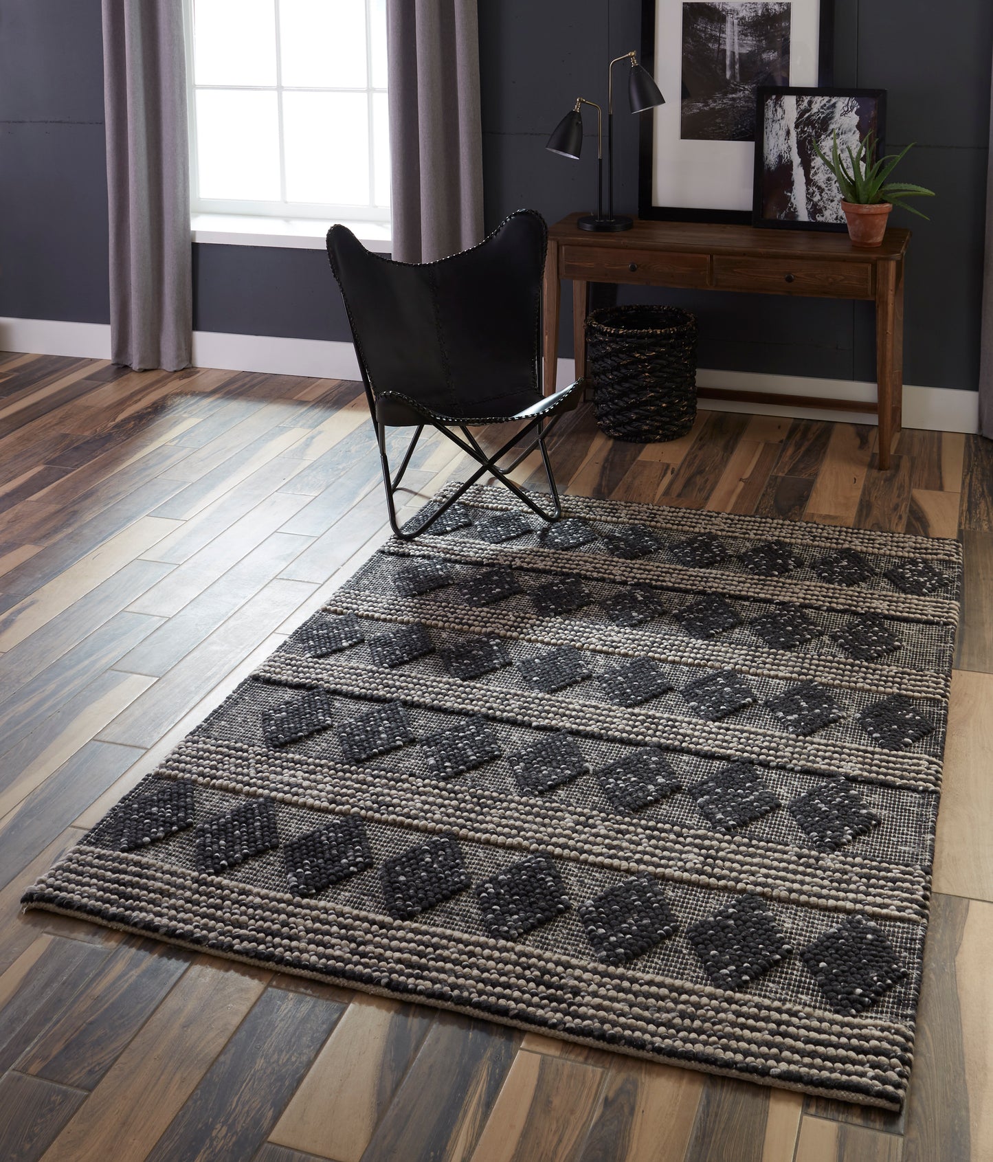 Dark gray geometric woven area rug with diamond motifs on wood floor, paired with a black butterfly chair and a wooden desk.