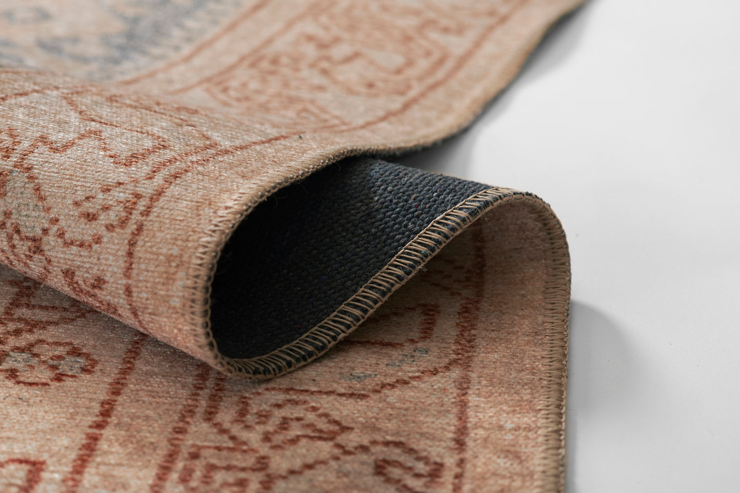 Peach-beige rug with red damask pattern, rolled to show dark navy underside and exposed whipstitch edge.