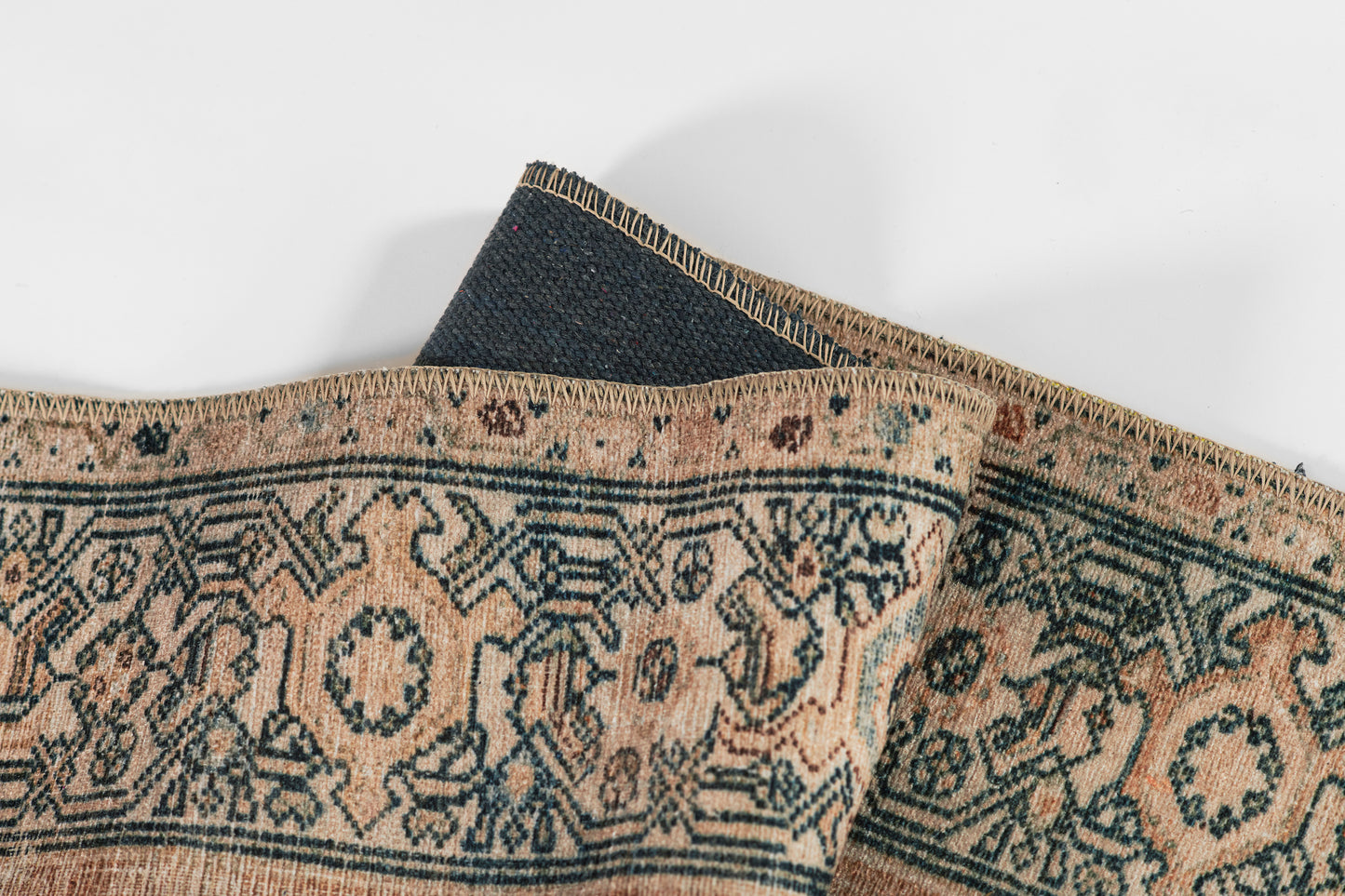 Beige oriental rug with dark blue geometric medallions and teal accents; zigzag-stitched border, folded corner.