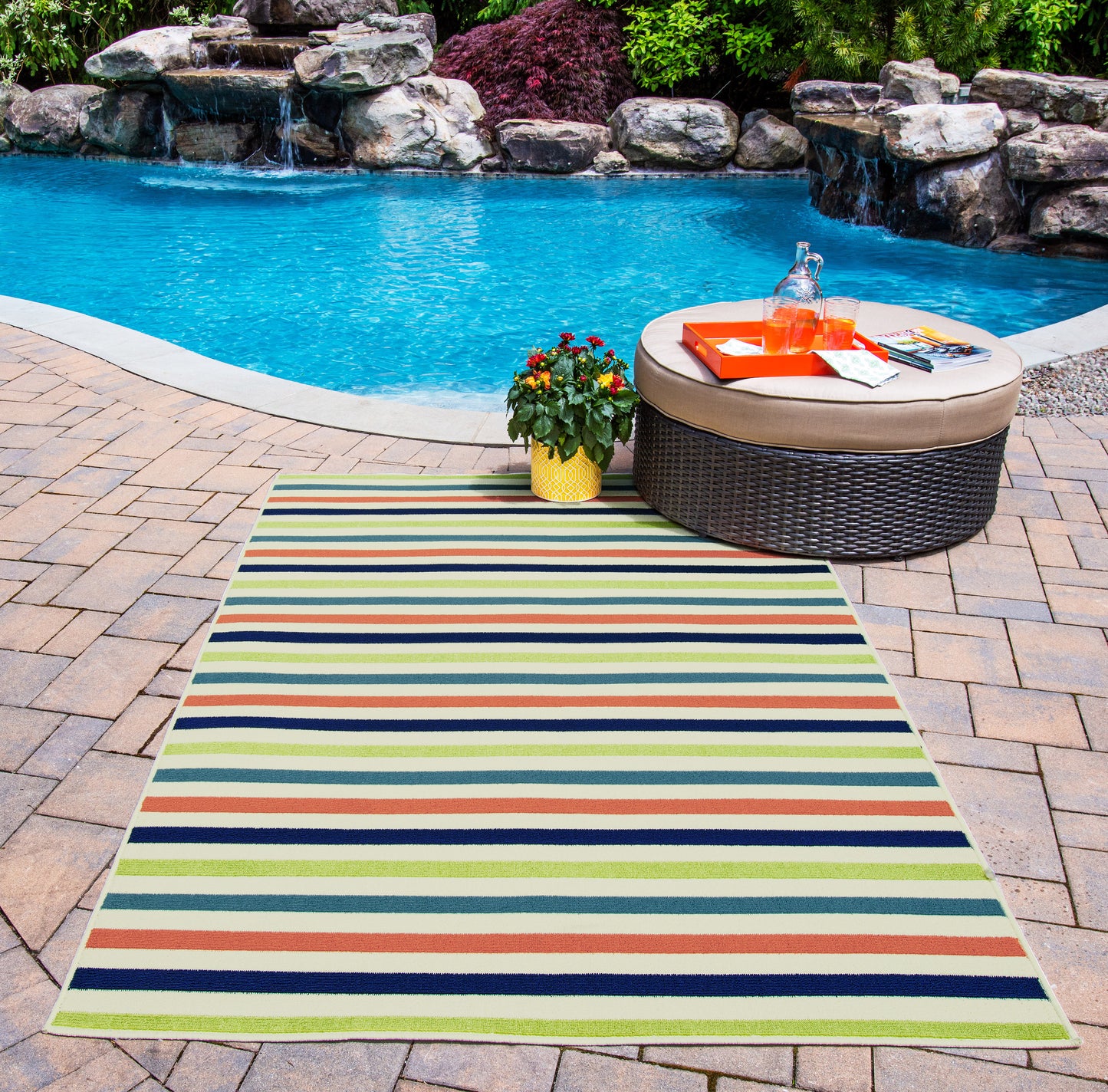Colorful outdoor striped rug on a brick pool deck beside a wicker ottoman with an orange tray and a potted plant.