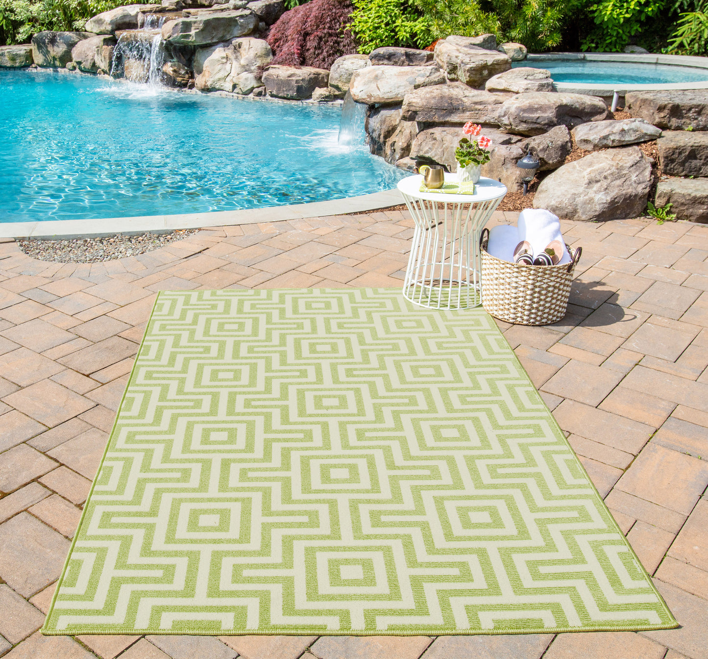 Green geometric outdoor rug on brick patio beside a white wire table & wicker basket by pool.
