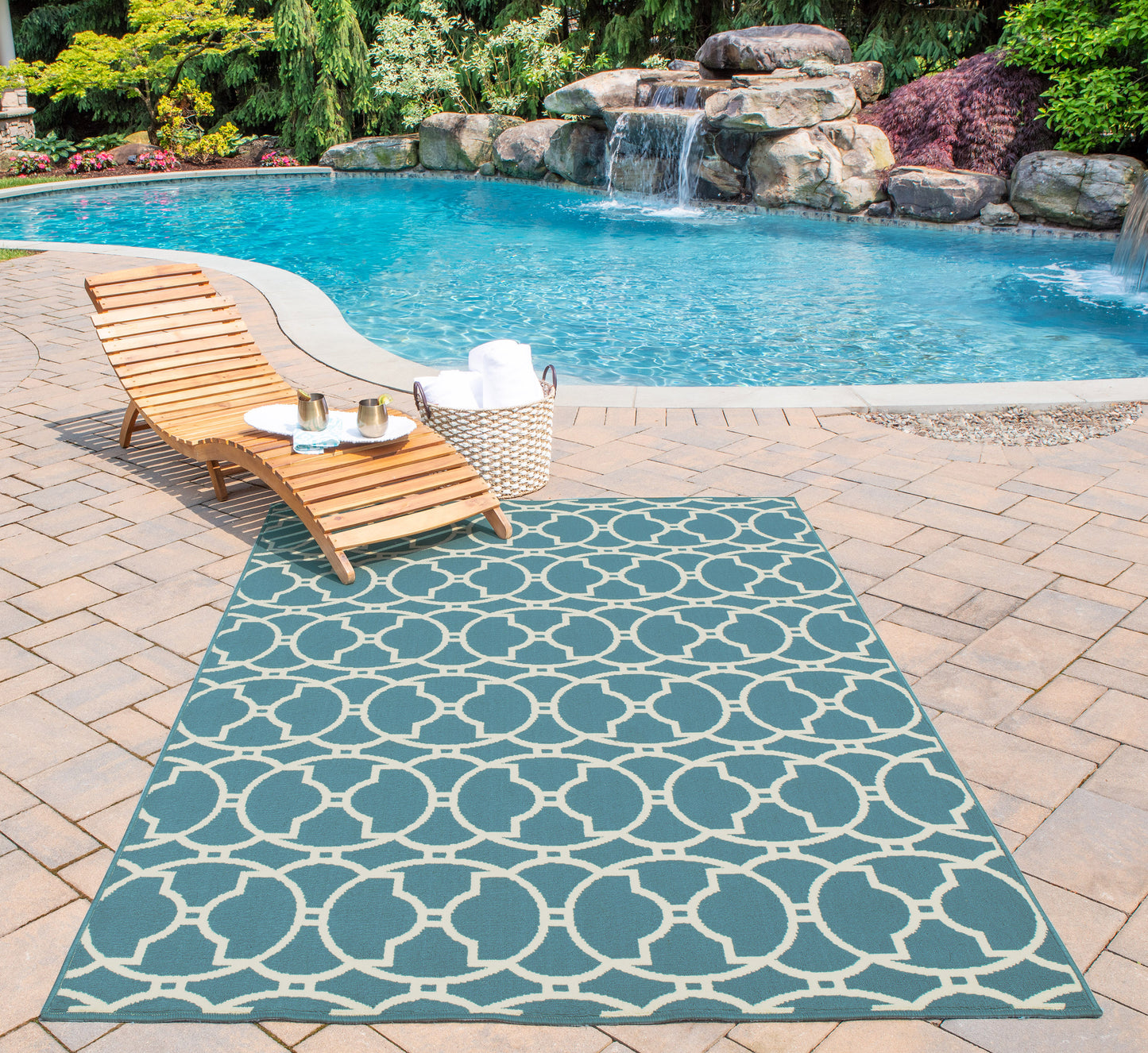 Blue Baja outdoor rug with white circular lattice pattern on brick pool deck beside a wooden lounge chair.