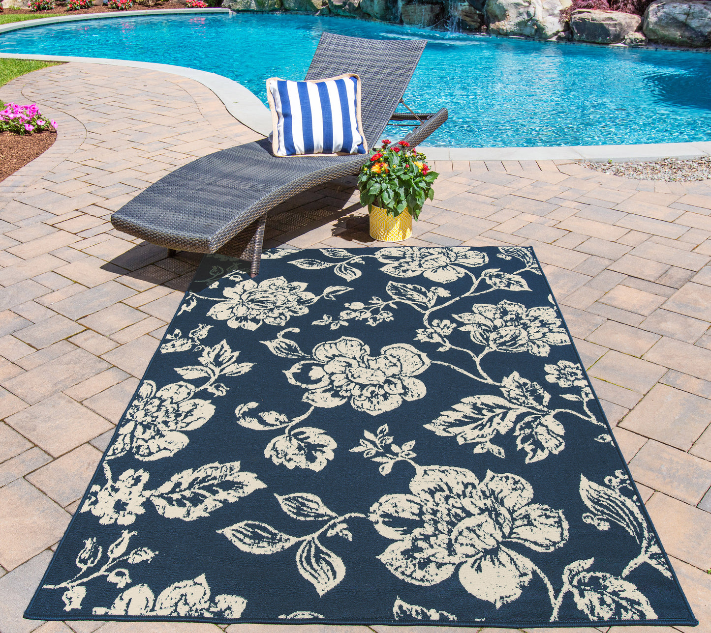 Poolside scene with navy floral outdoor rug, wicker lounge chair, blue & white striped pillow, potted plant on brick patio.
