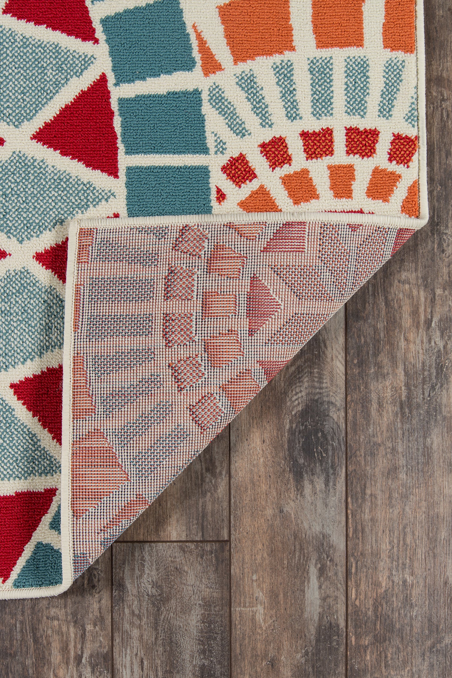 Colorful geometric rug on wood floor; bold red, teal, orange blocks with cream accents; folded corner shows pink backing.
