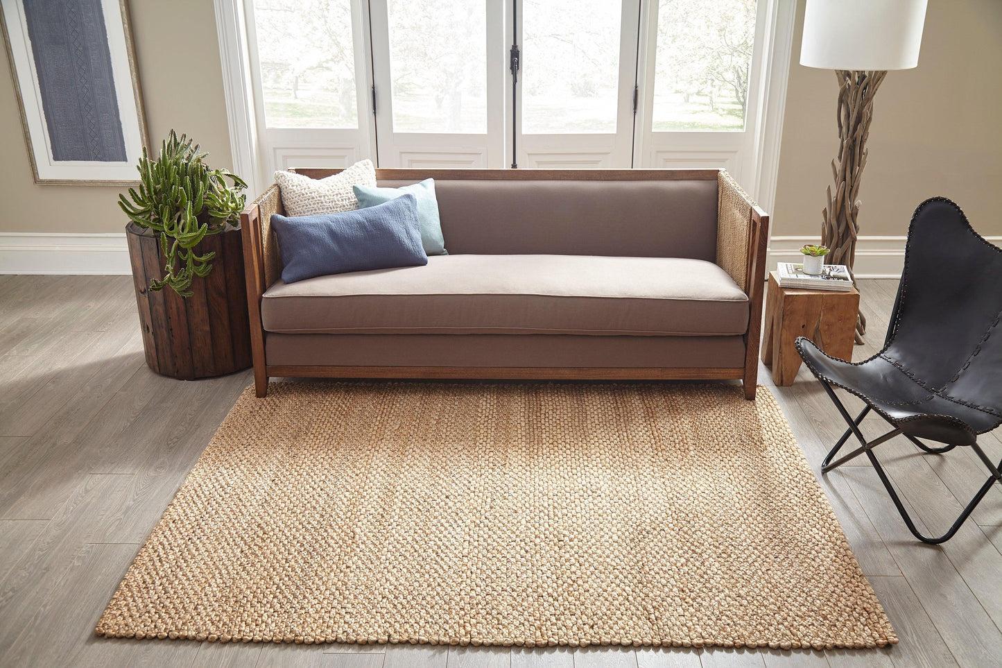 Natural jute area rug under wood-frame sofa with blue and cream pillows, black leather butterfly chair, and potted plant.
